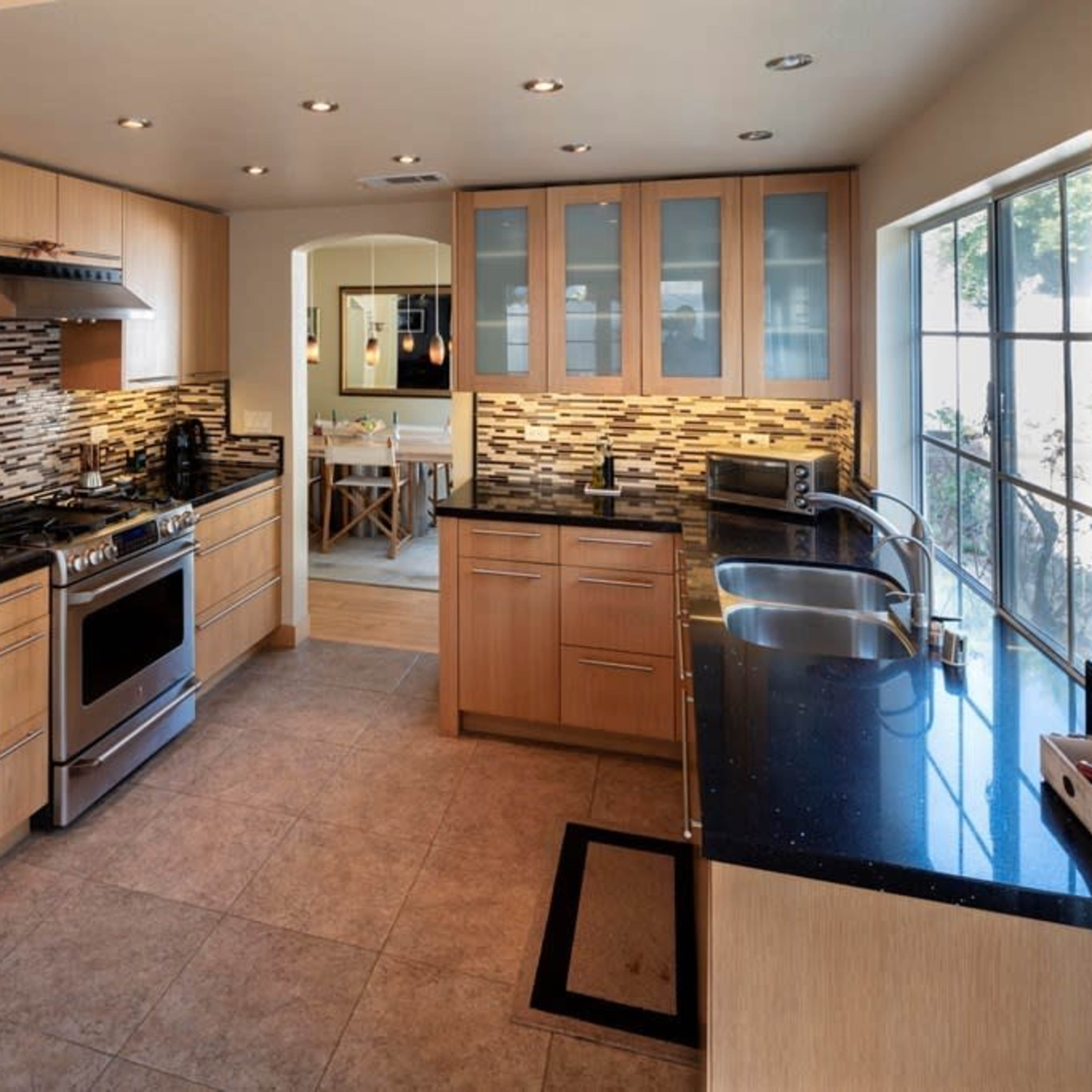 A modern kitchen with wooden cabinetry, black countertops, a stainless steel stove, and a window overlooking a dining area.