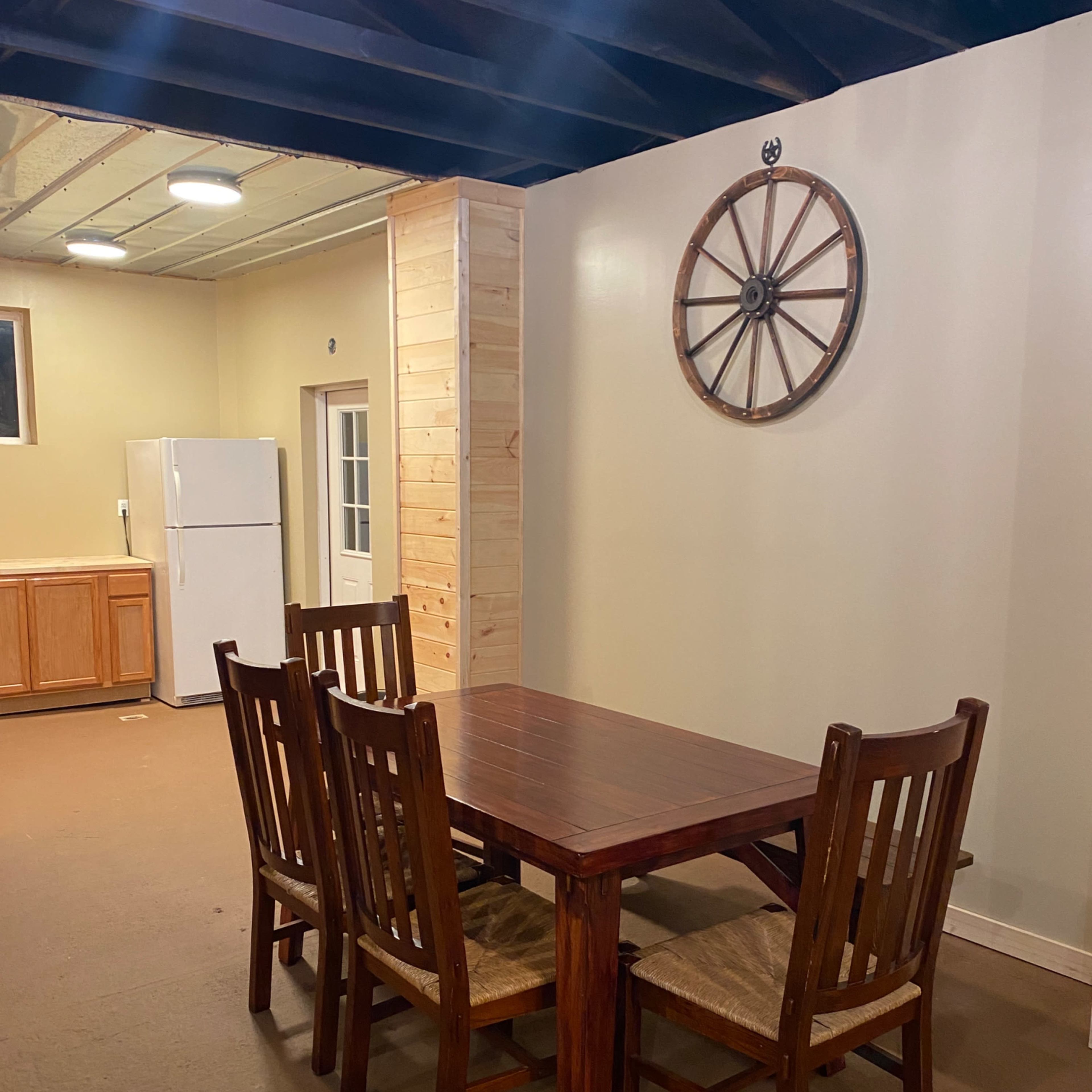 A wooden dining table with four chairs is set in a spacious room featuring a wheel decoration on the wall and a refrigerator in the background.