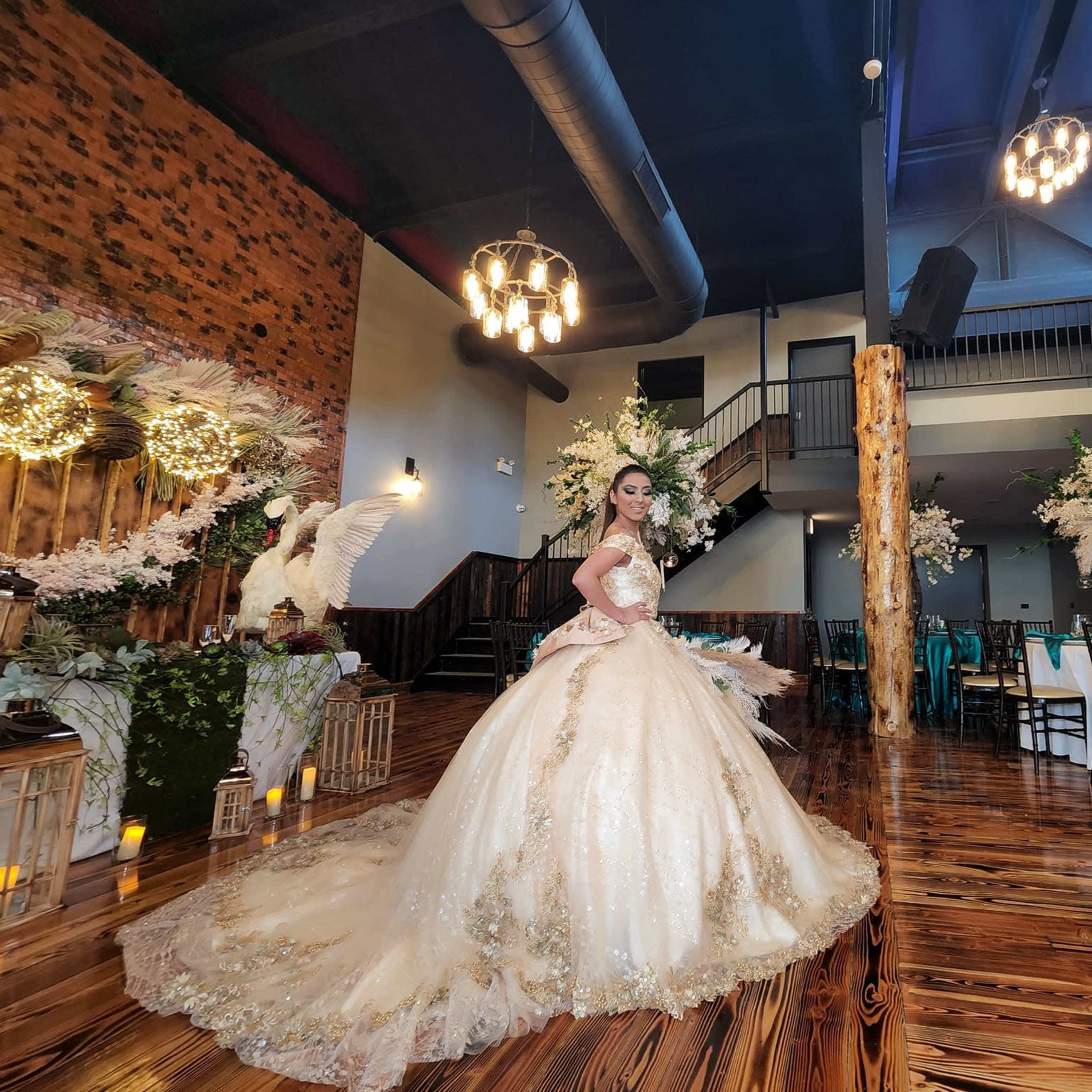 A bride in a golden wedding gown poses in a decorated reception venue with floral arrangements and elegant lighting.