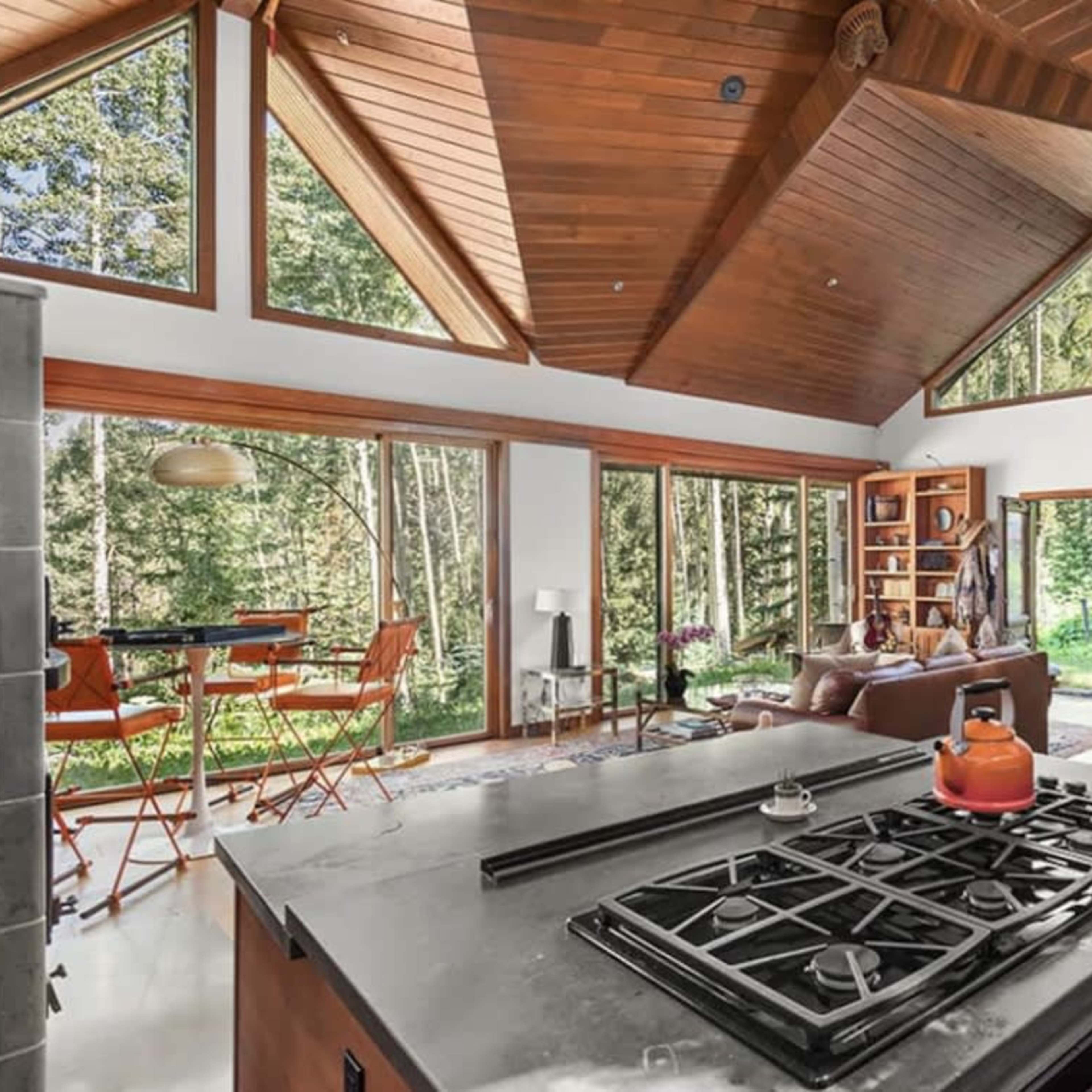 A modern kitchen with a central island, featuring a gas stove and stainless steel appliances, alongside large windows that offer views of a wooded area.