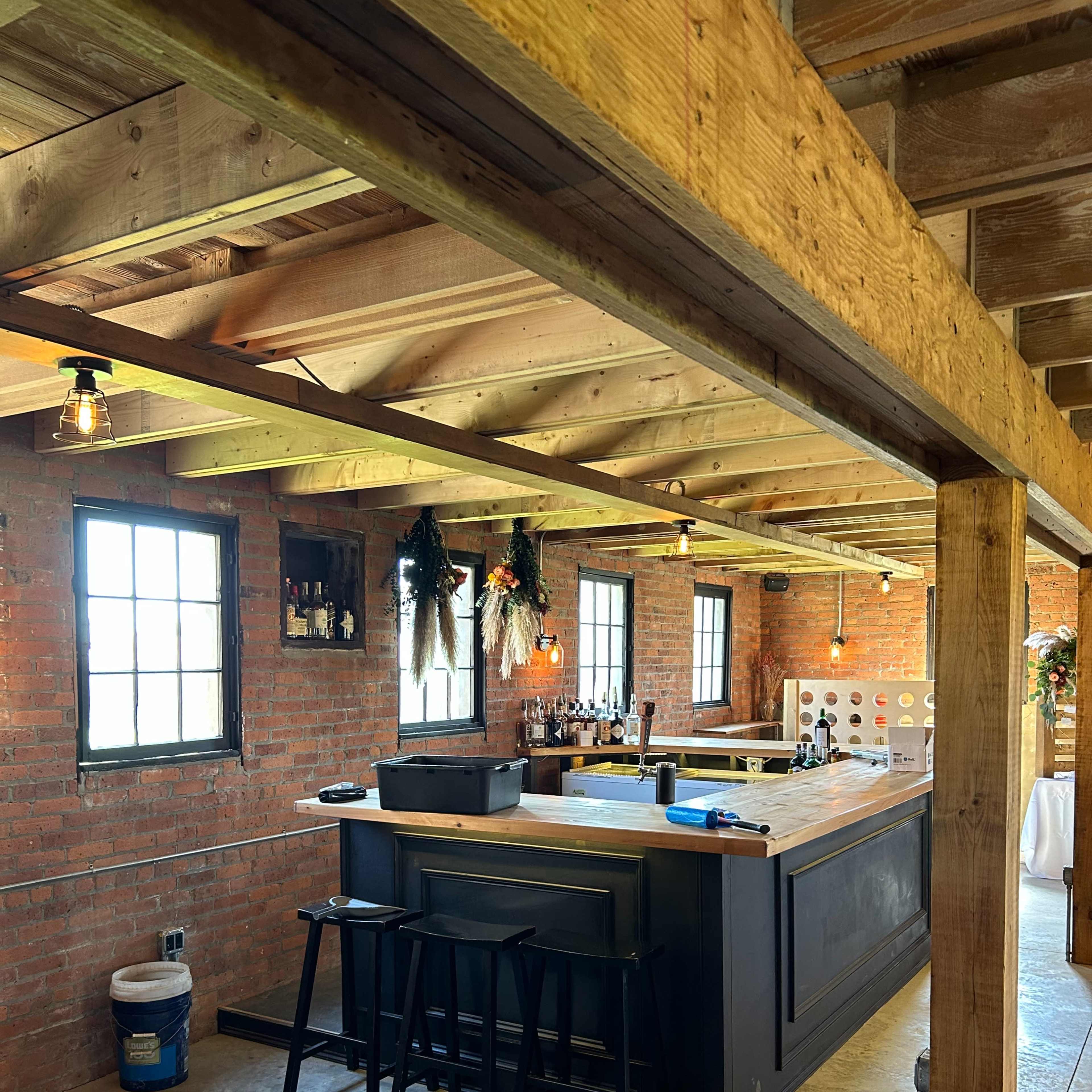 A rustic bar area with a wooden counter, black stools, and exposed brick walls beneath a wooden ceiling.