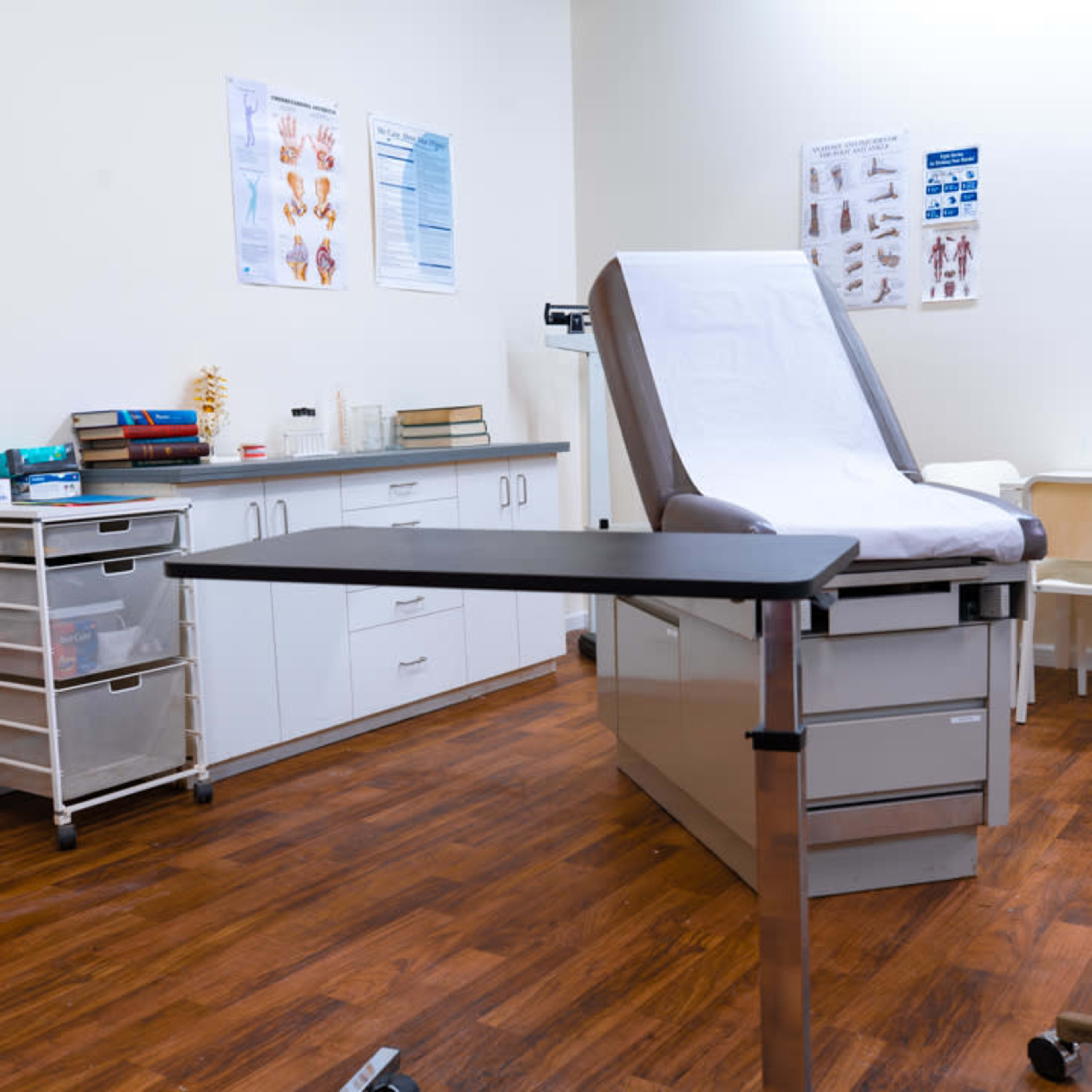A medical examination room features an examination table, a cart with supplies, and a small table with chairs.