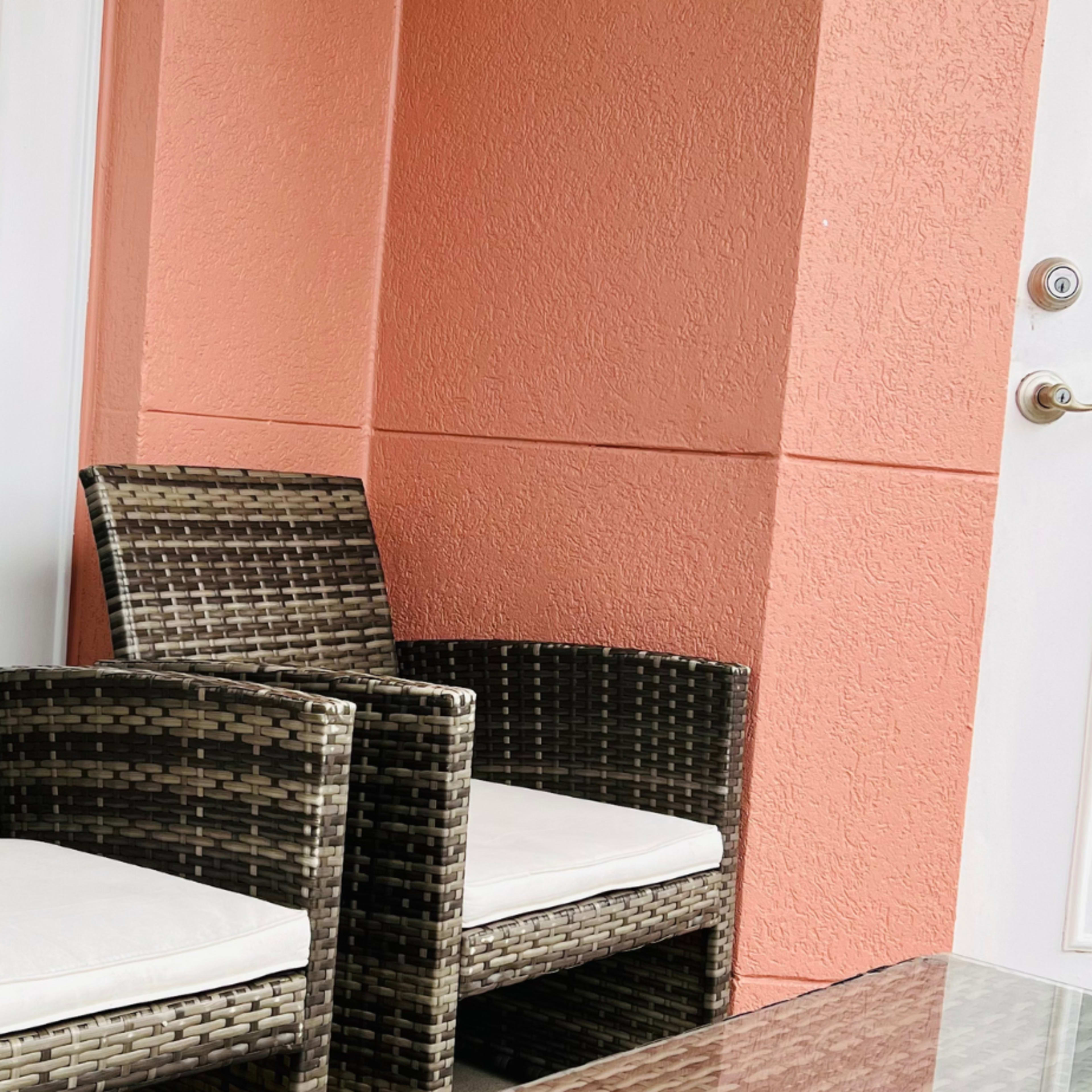 Two wicker chairs with cushions are positioned beside a glass table against a peach-colored wall and a window.