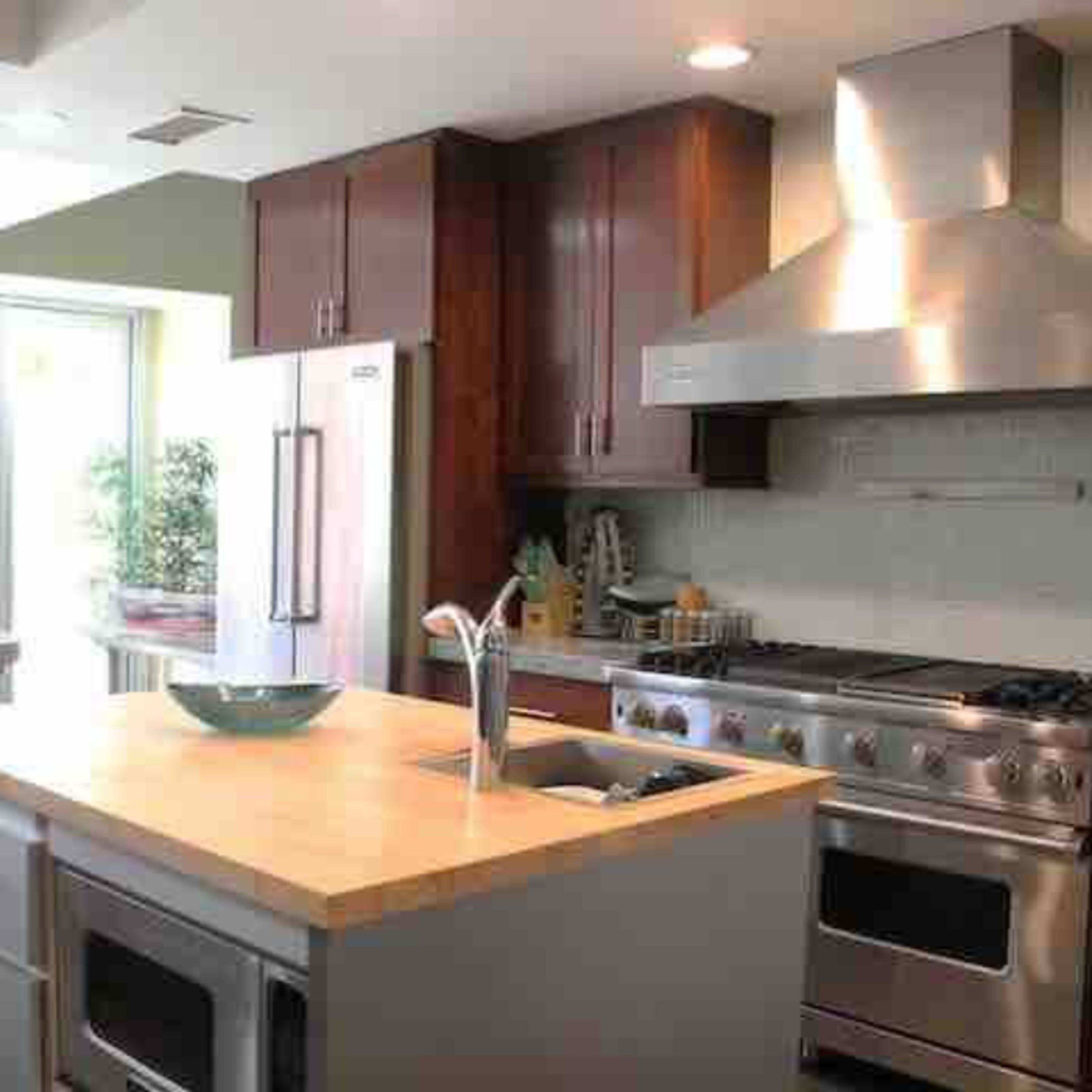 A modern kitchen with wooden cabinets, a large island featuring a wooden countertop, and a stainless steel range hood above a gas stove.
