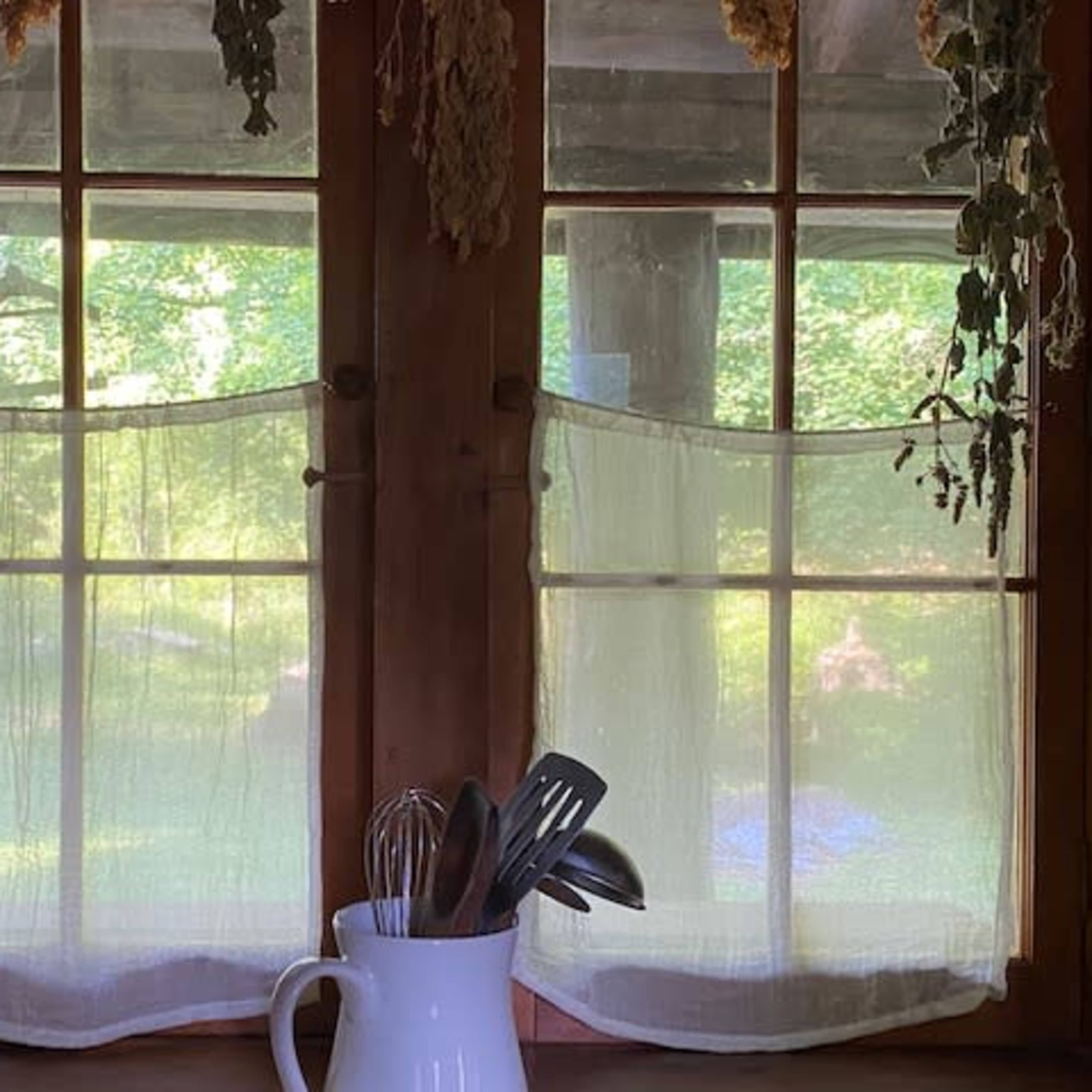 A window with sheer curtains, a white pitcher, and kitchen utensils on a windowsill, with green foliage visible outside.