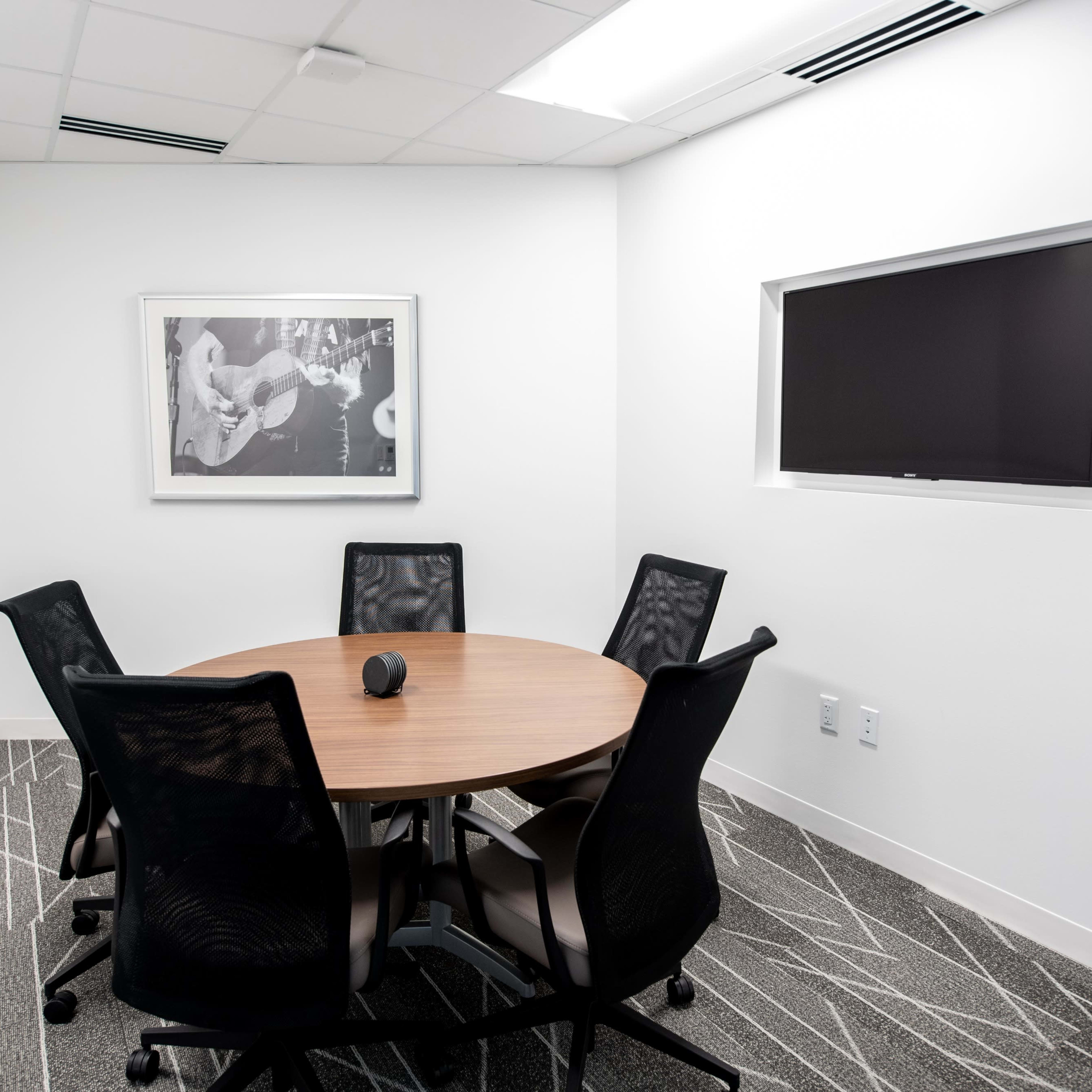 A small meeting room features a round wooden table surrounded by five black mesh chairs, with a framed photo on the wall and a mounted screen.