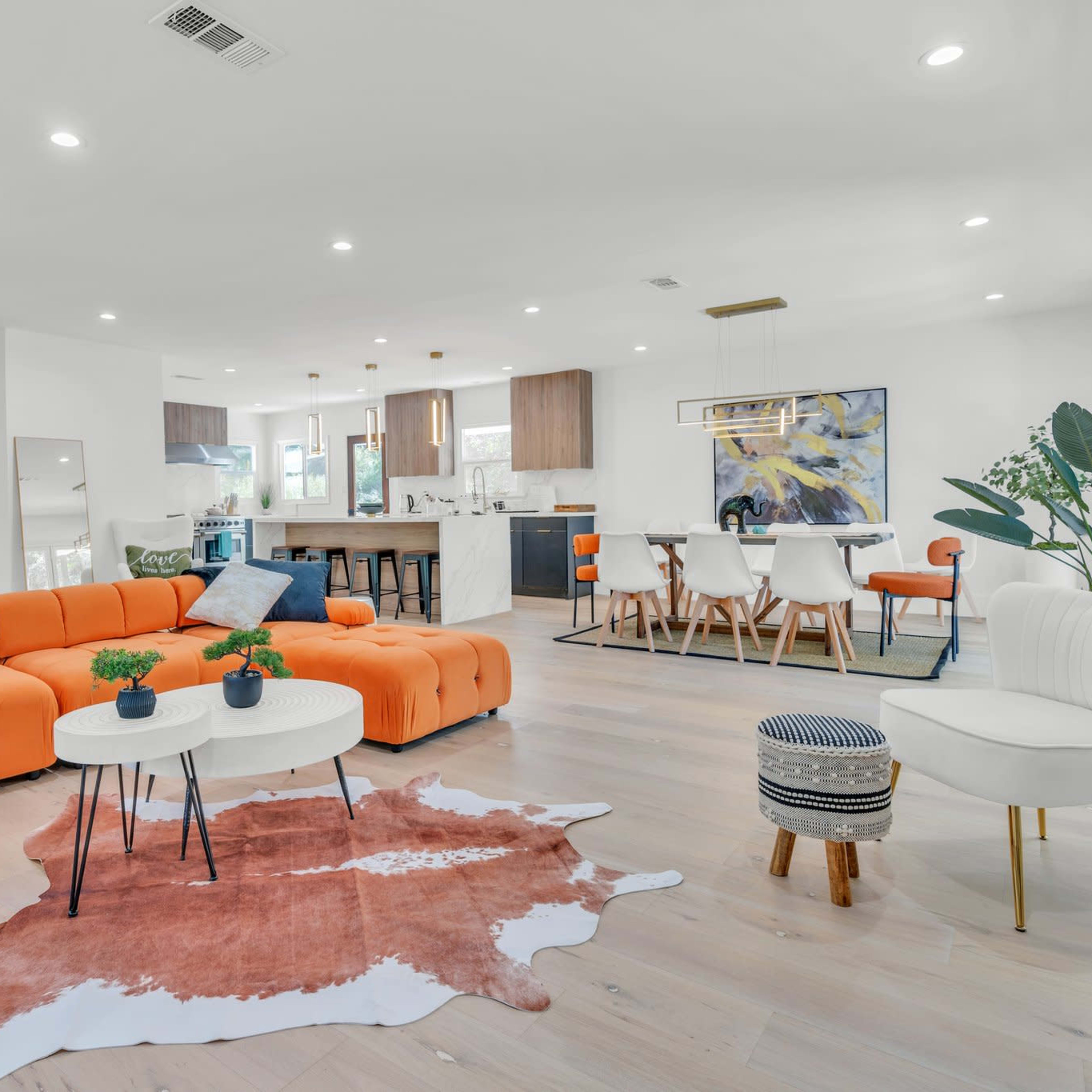 A modern living area features an orange sectional sofa, a round coffee table, and a dining space with a wooden table, surrounded by white walls and wooden flooring.