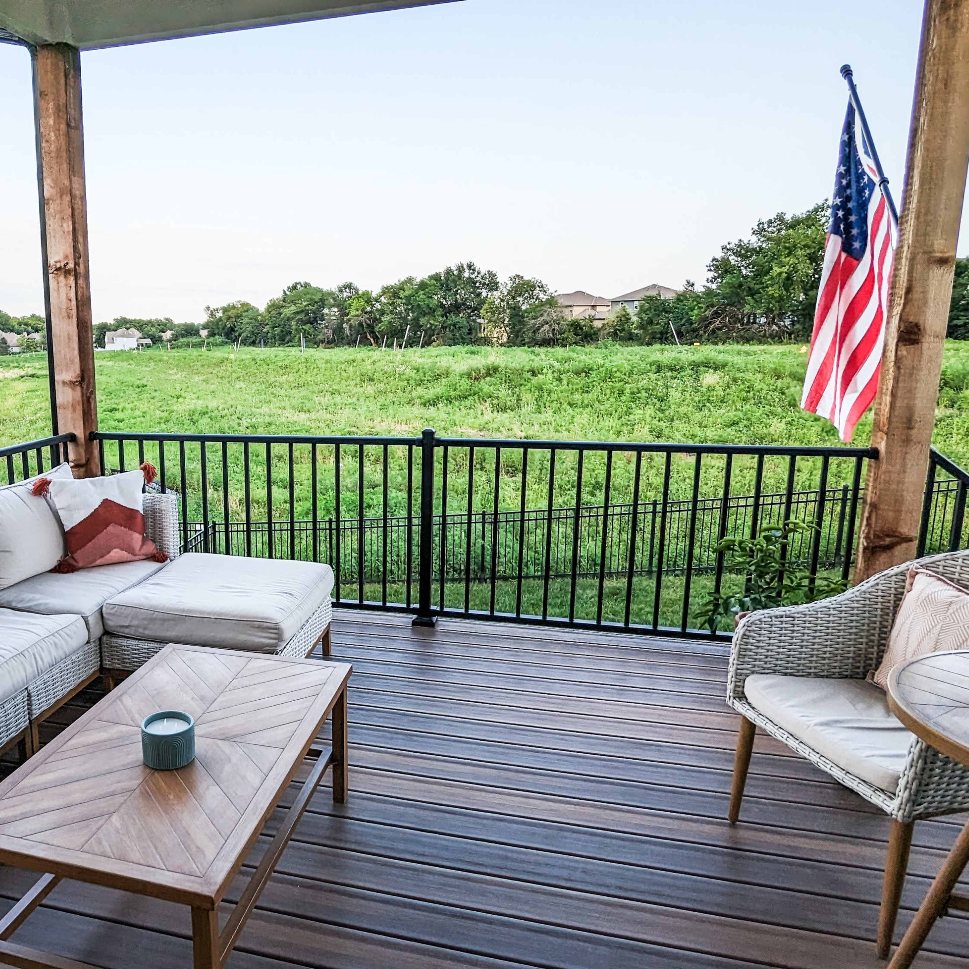 A covered patio features a sectional sofa, a wooden coffee table, wicker chairs, and an American flag, with a green field and trees visible in the background.