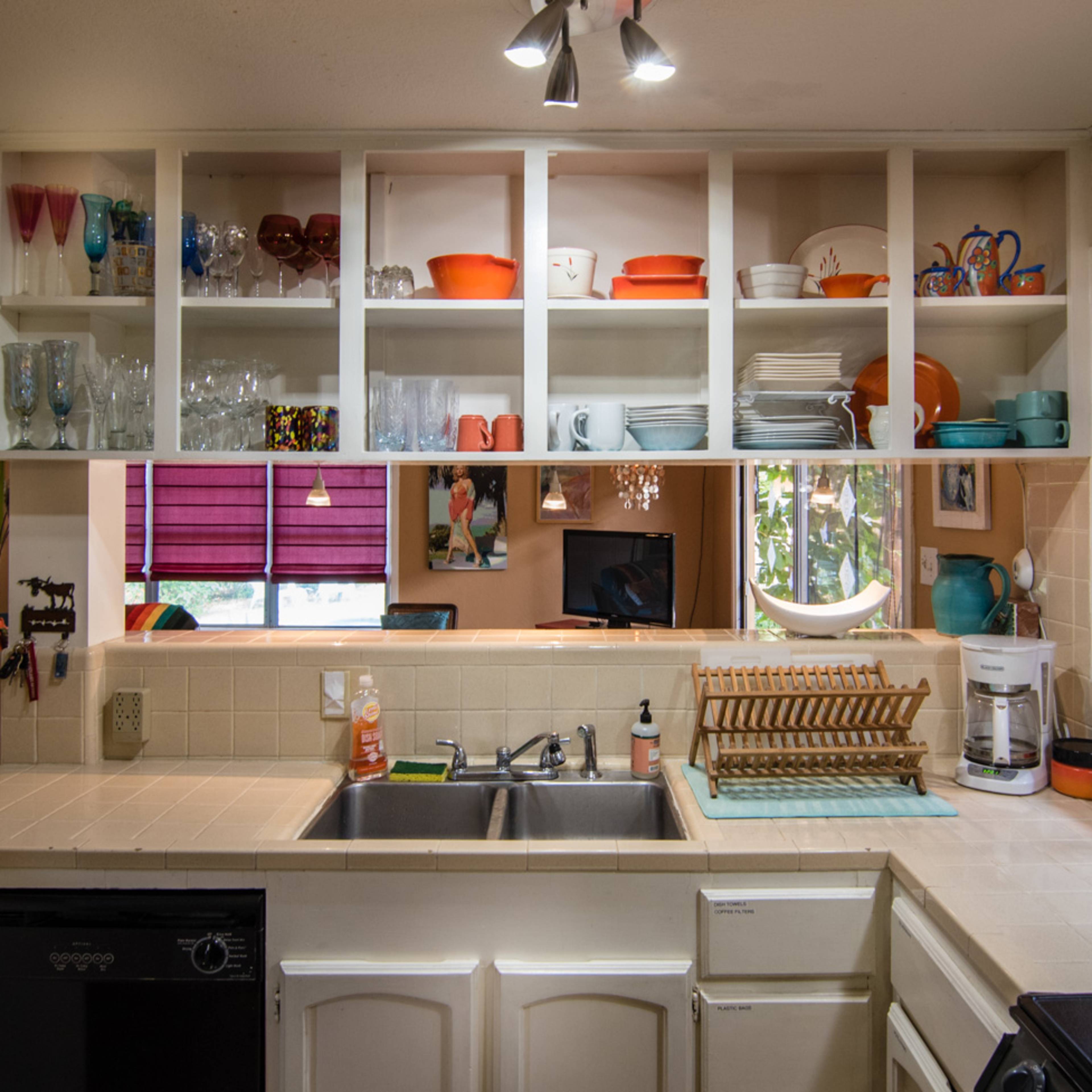 A kitchen with white tiled countertops, a double sink, and open shelves displaying colorful dishes and glassware.