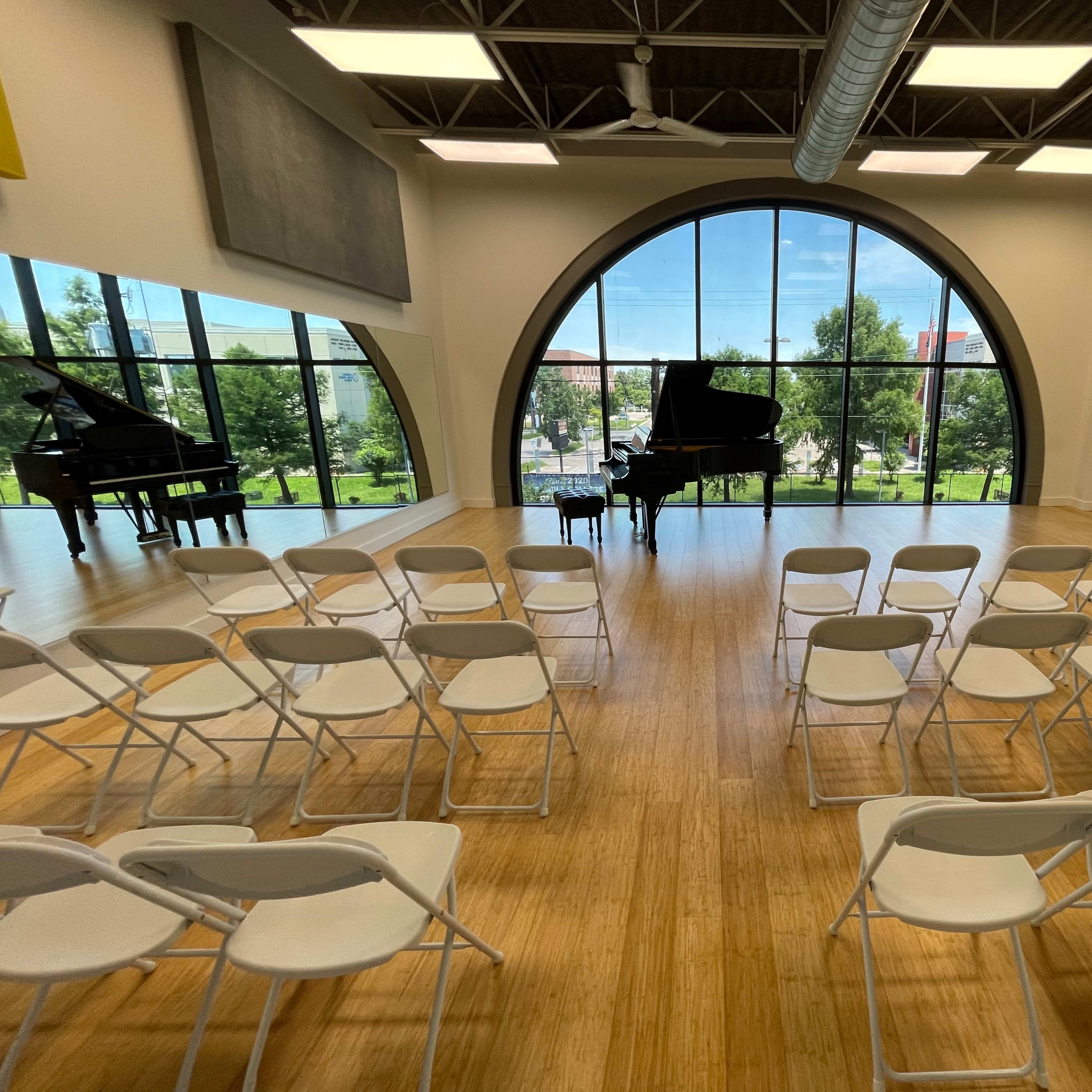 A spacious music room with two grand pianos and white folding chairs arranged in front of them, set against large windows that overlook a green landscape.
