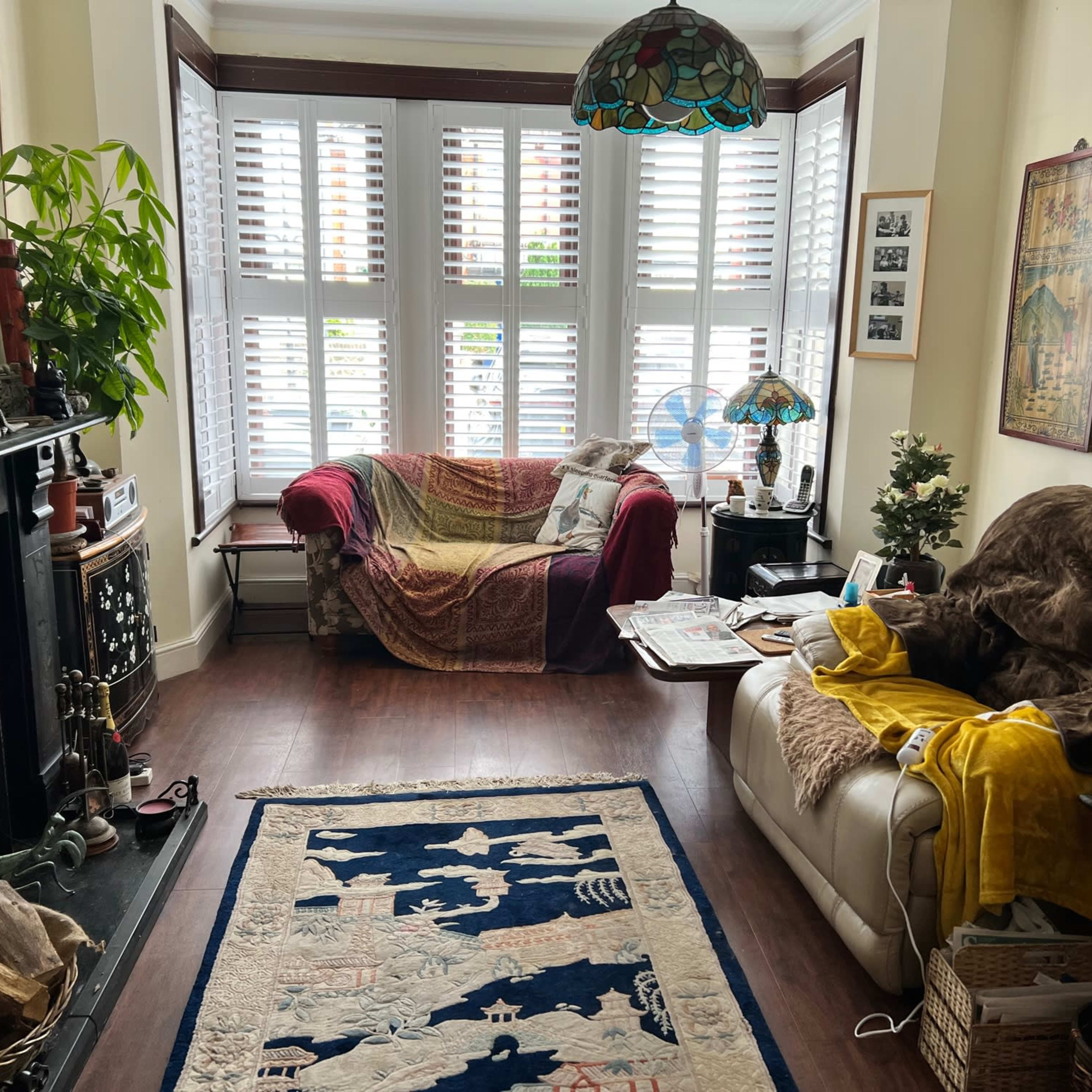 A cozy living room features a red couch, a white sofa covered in yellow and brown blankets, a patterned rug, and plants beside a decorative fireplace.