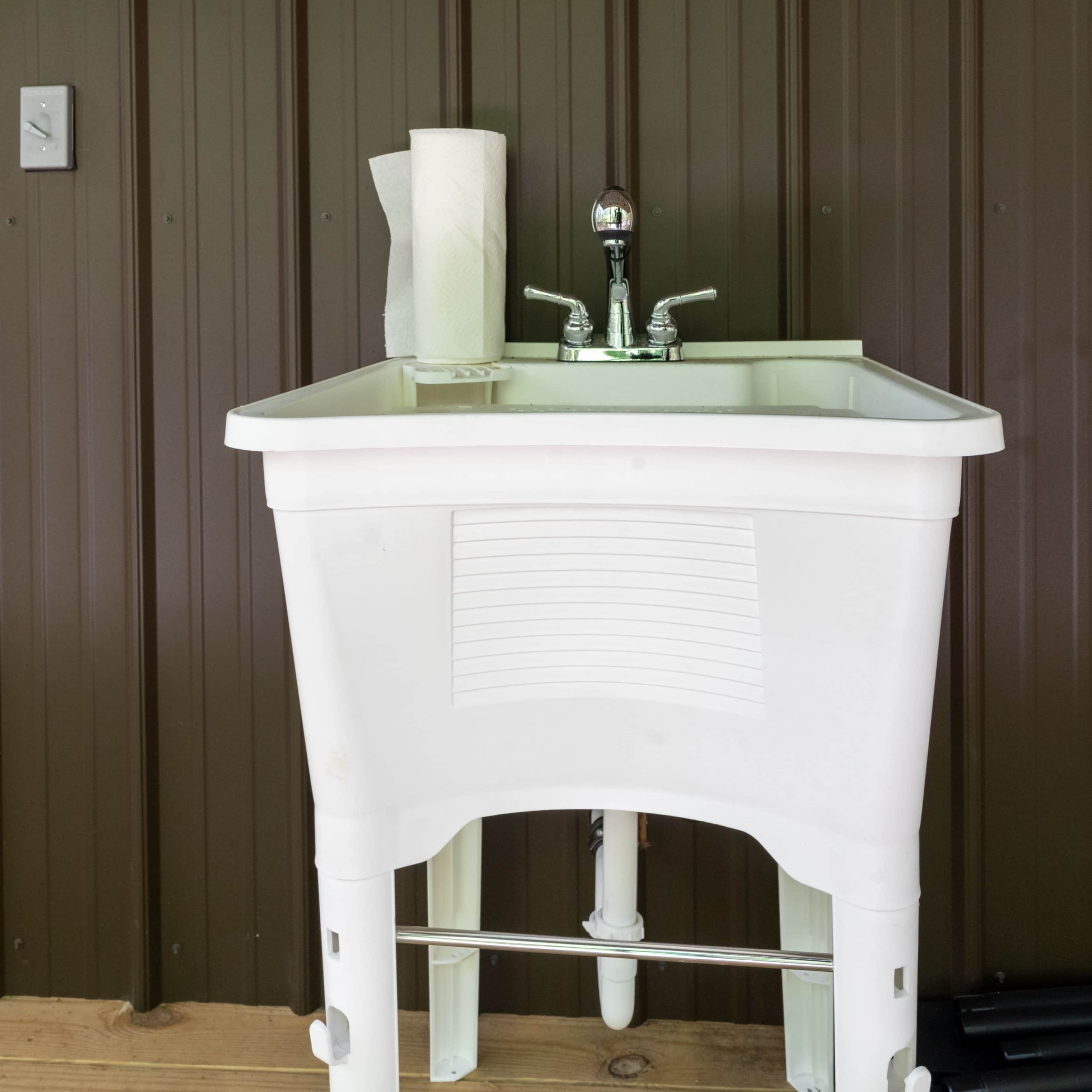 A white utility sink is mounted against a brown corrugated wall, with paper towels positioned beside the faucet.