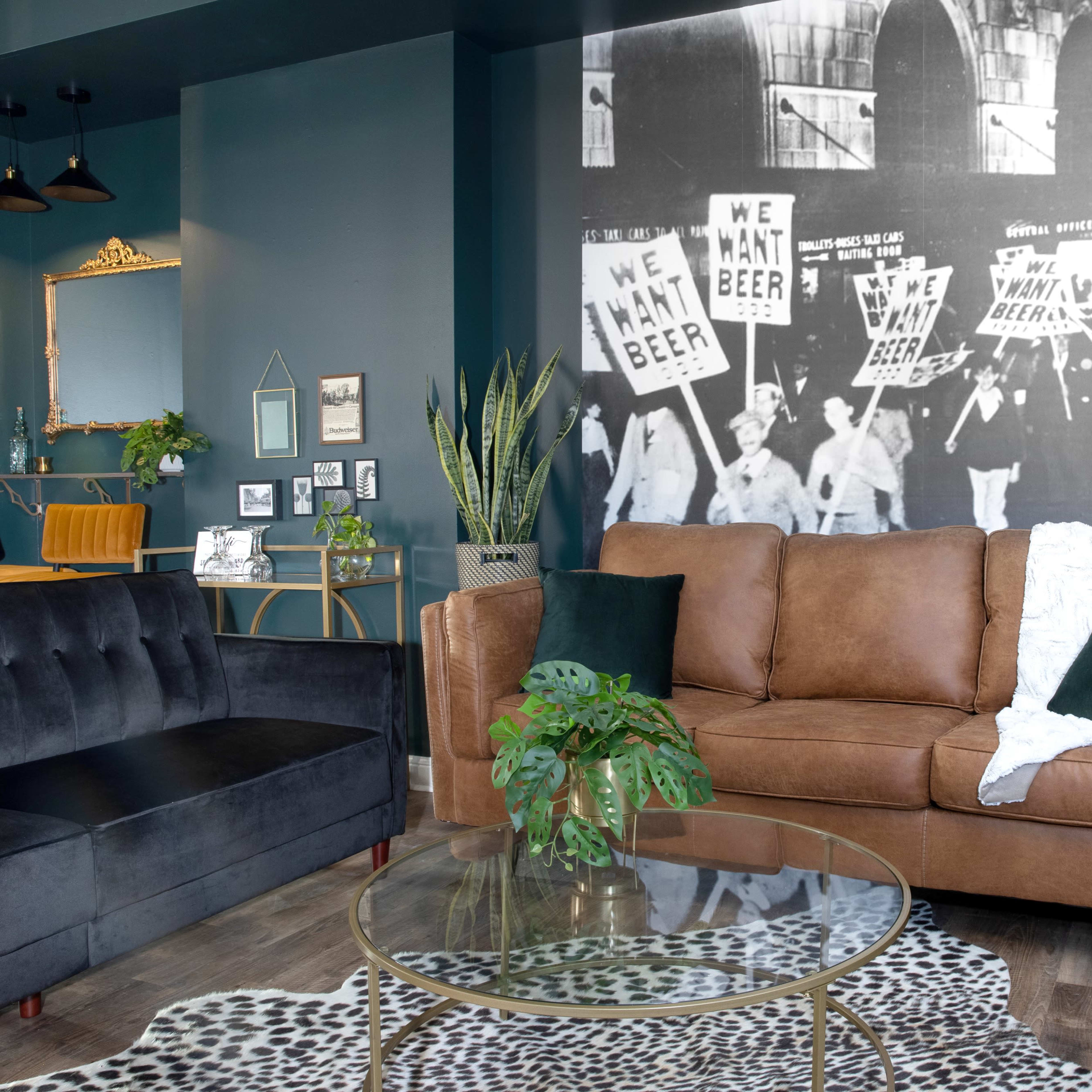 A living room with a brown leather sofa, a glass coffee table on a leopard print rug, and a large black-and-white mural depicting a historical protest for beer.