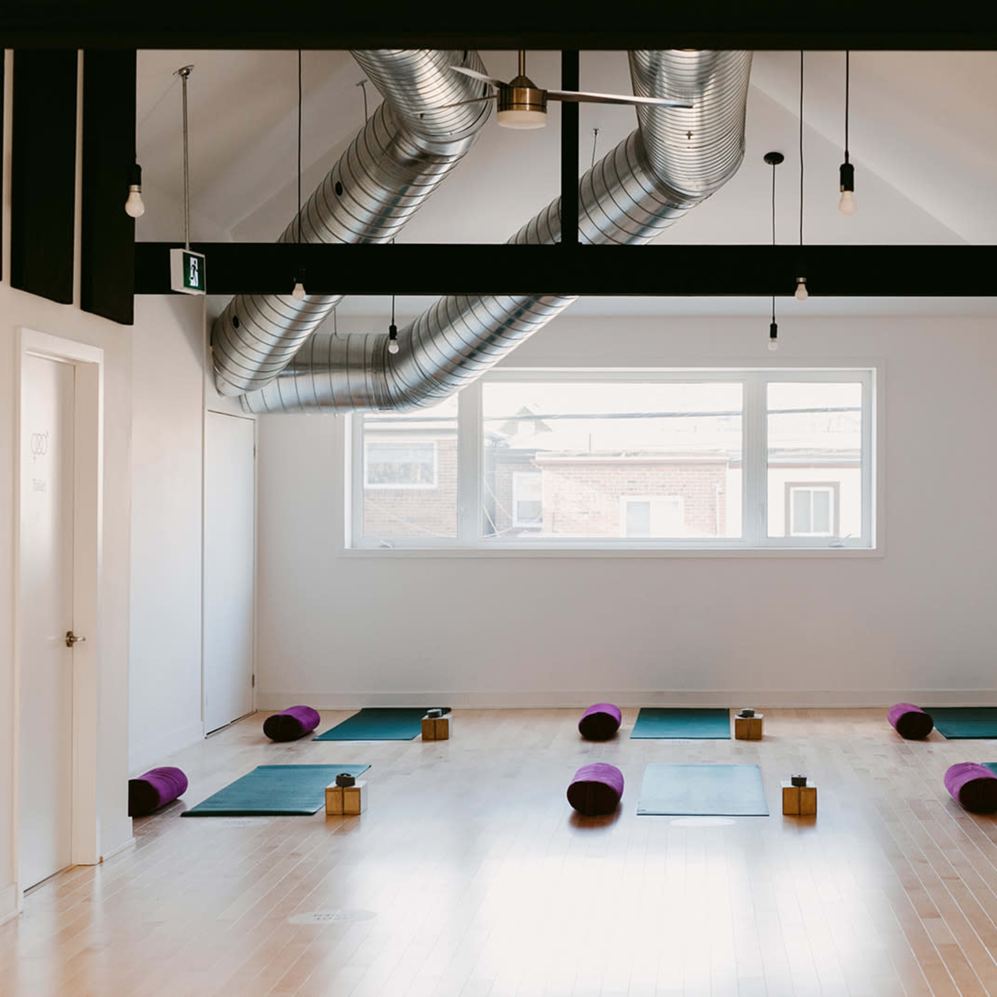A spacious yoga studio with mats and purple bolsters arranged neatly on the wooden floor under exposed ductwork and minimal lighting.