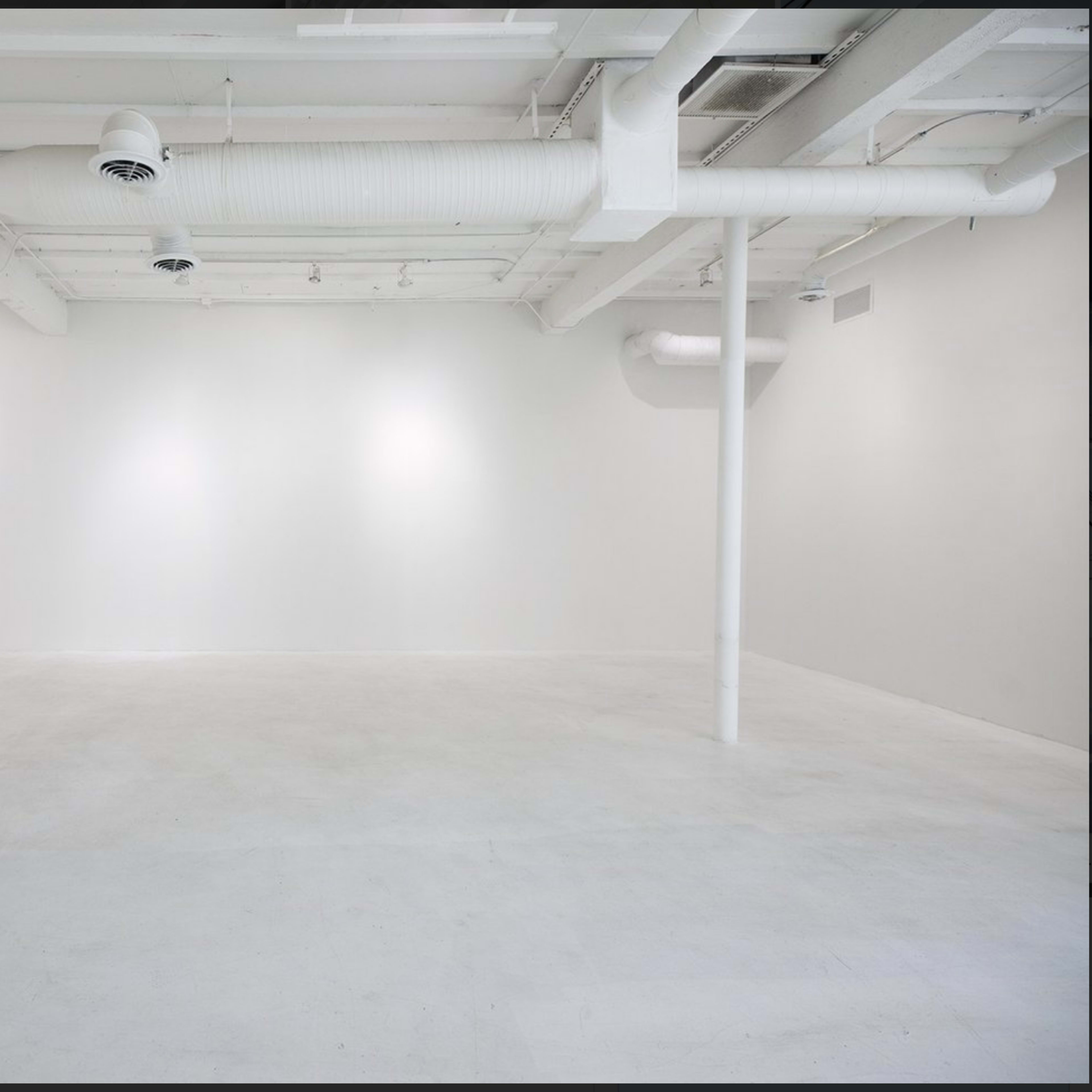 A spacious, empty room with white walls and a white floor, featuring several columns and overhead ventilation ducts.