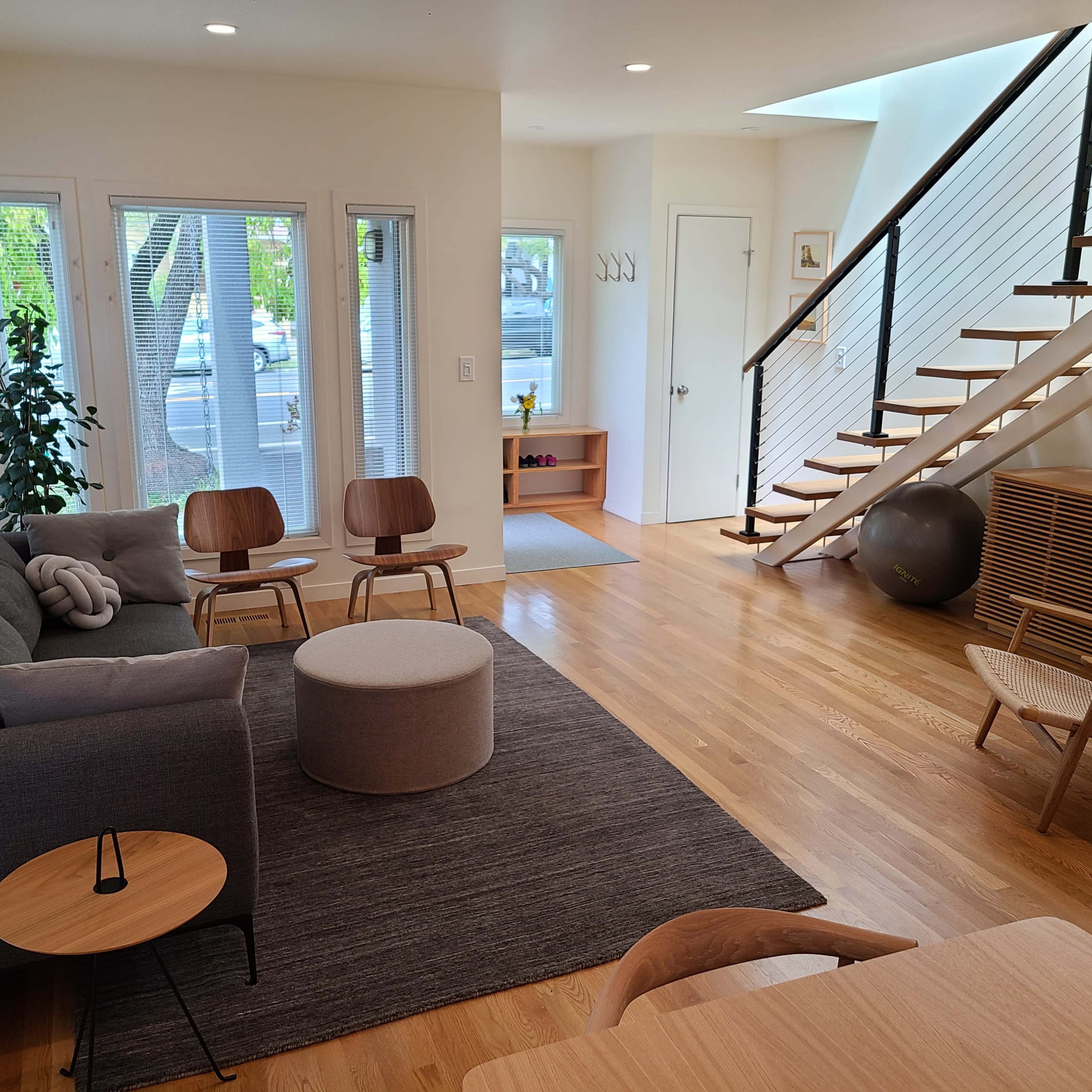 A modern living room featuring a gray sofa, two wooden chairs, a round ottoman, and a staircase with a wooden railing.