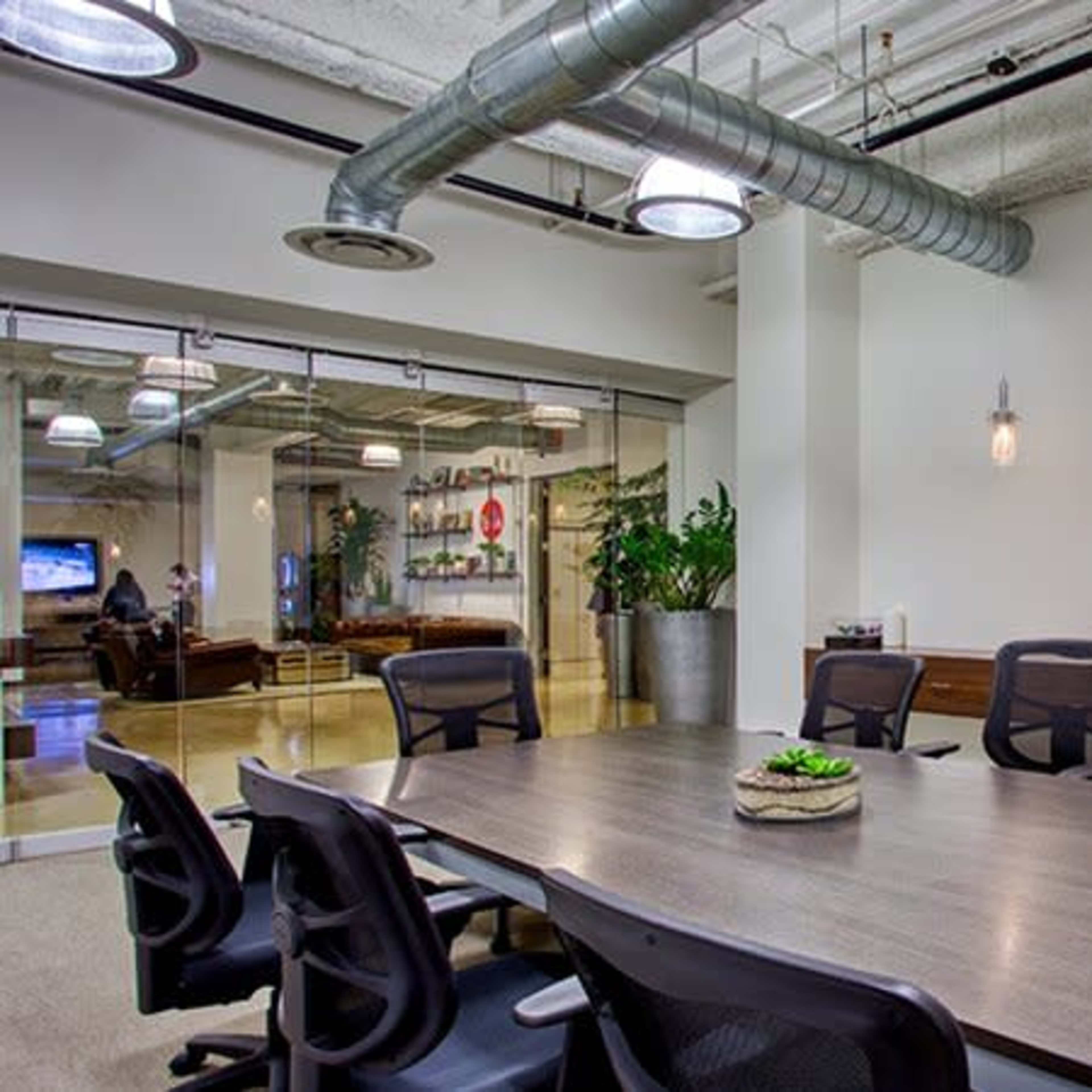 A modern conference room features a large wooden table surrounded by black chairs, with glass walls revealing a seating area beyond.