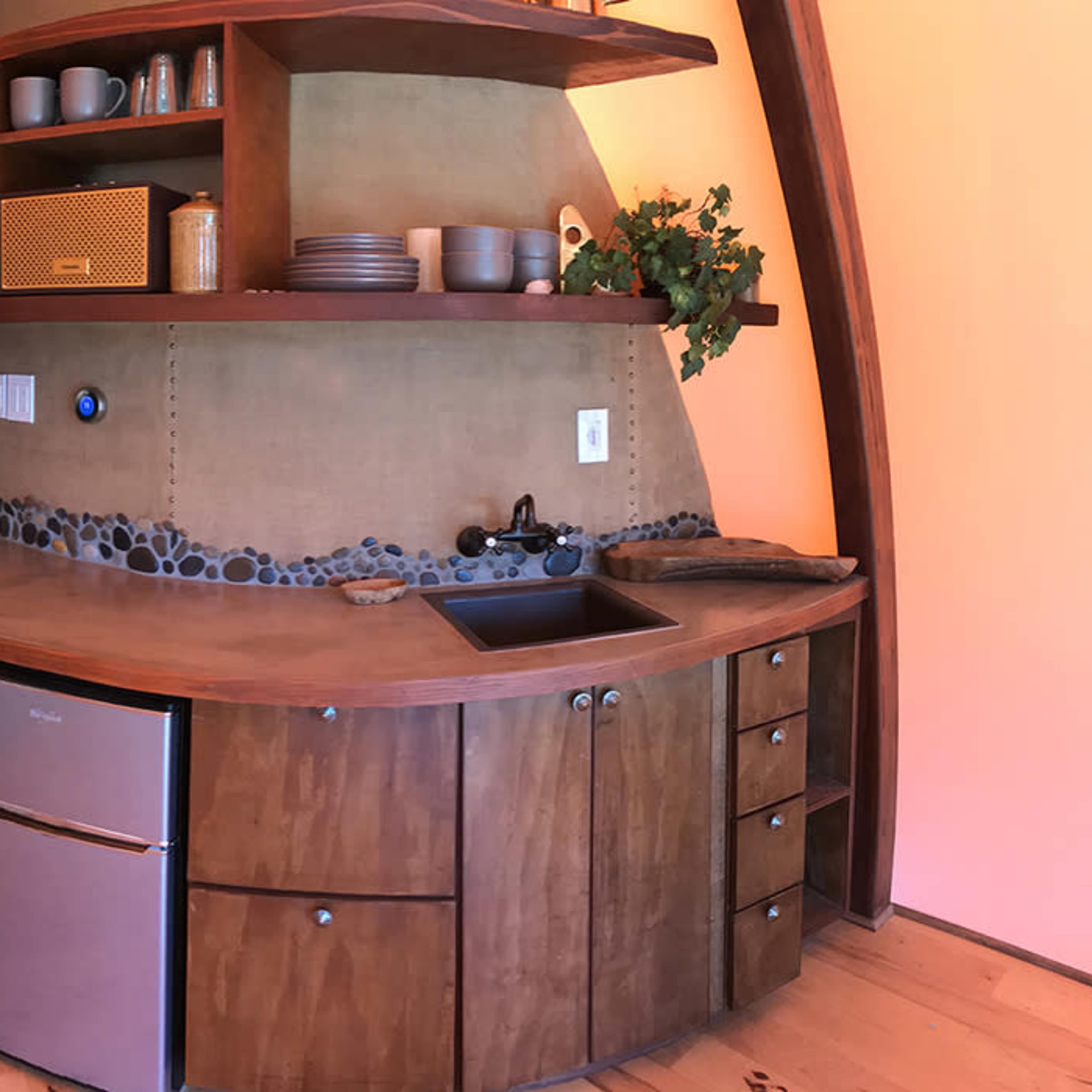 A wooden kitchen area with a curved countertop, sink, and a small refrigerator, backed by a wall of shelves holding dishes and decorative items.