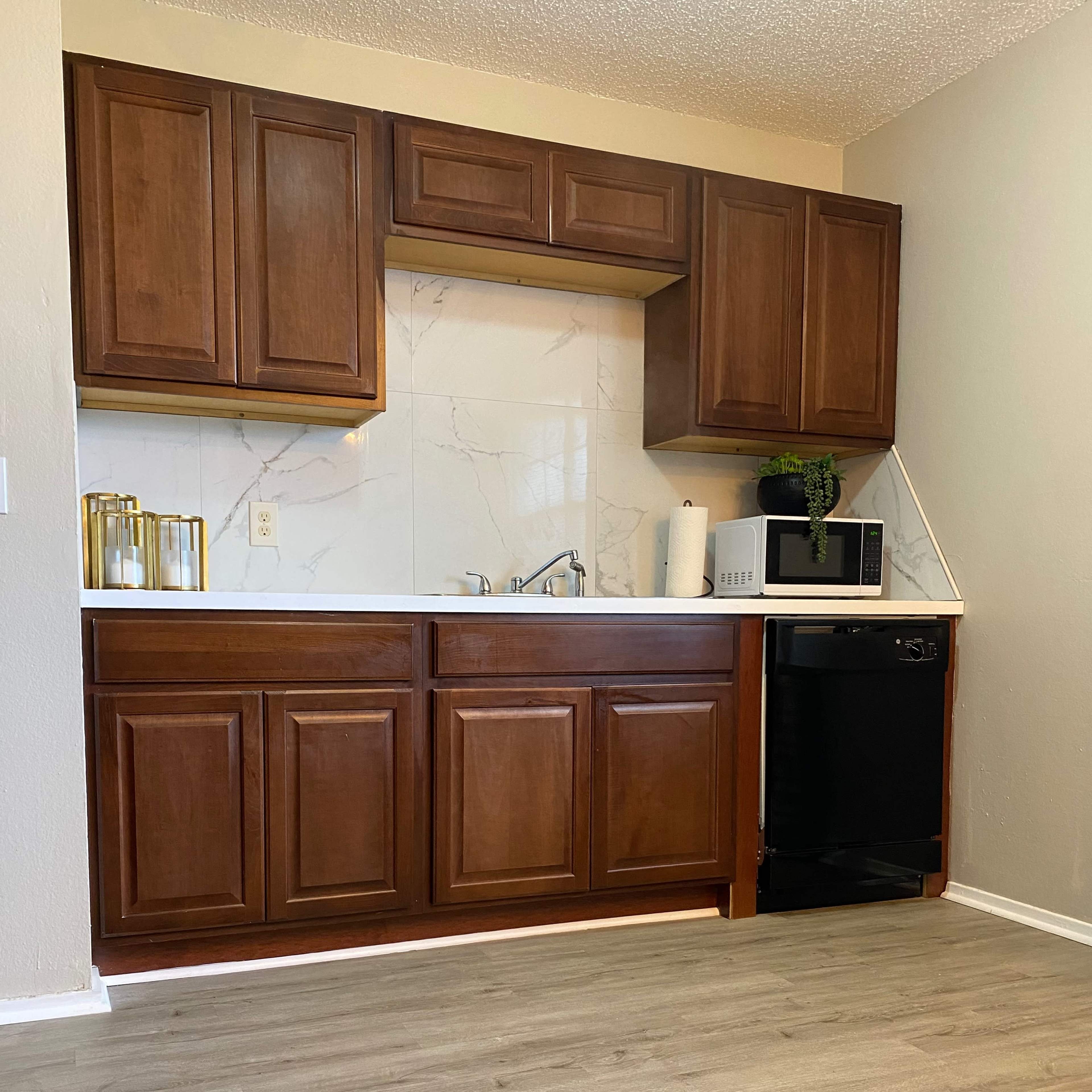 A small kitchen area featuring brown wooden cabinets, a white countertop, a sink, a microwave, and a black refrigerator, with light-colored laminate flooring.