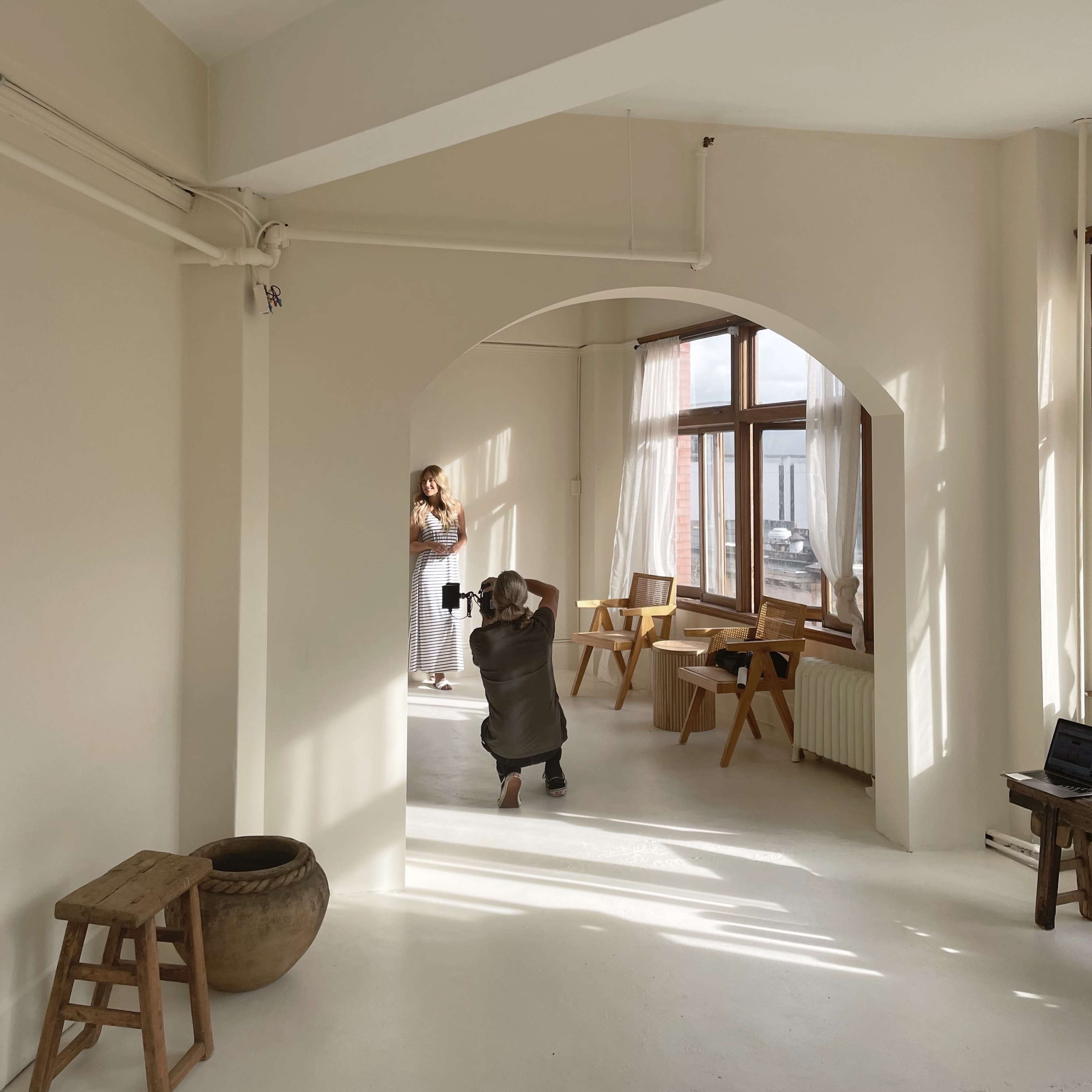 A photographer captures an image of a model in a bright, airy room with large windows and minimal furnishings.