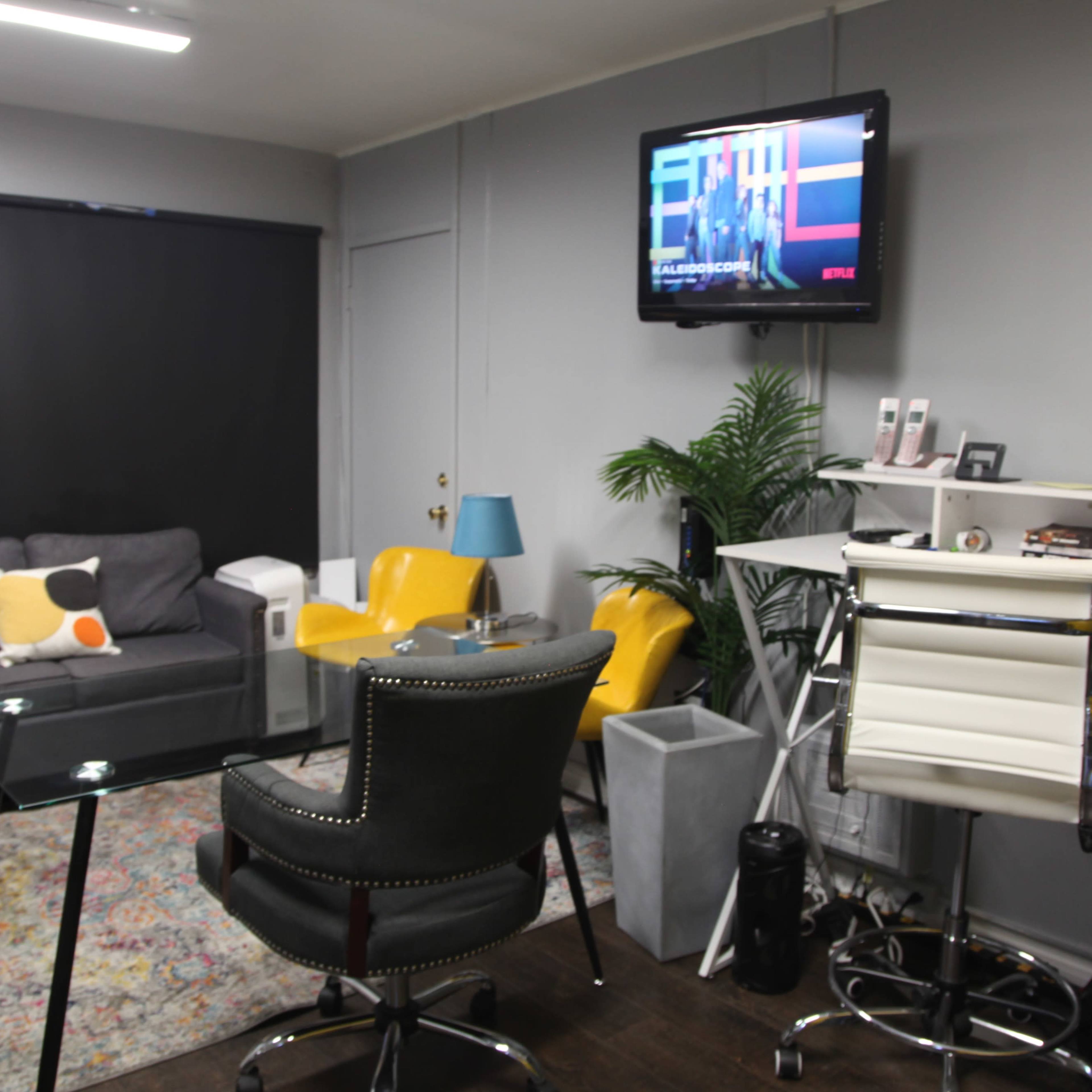 The room features a gray sofa, two yellow chairs, a glass table, a desk with a rolling chair, and a television mounted on the wall.