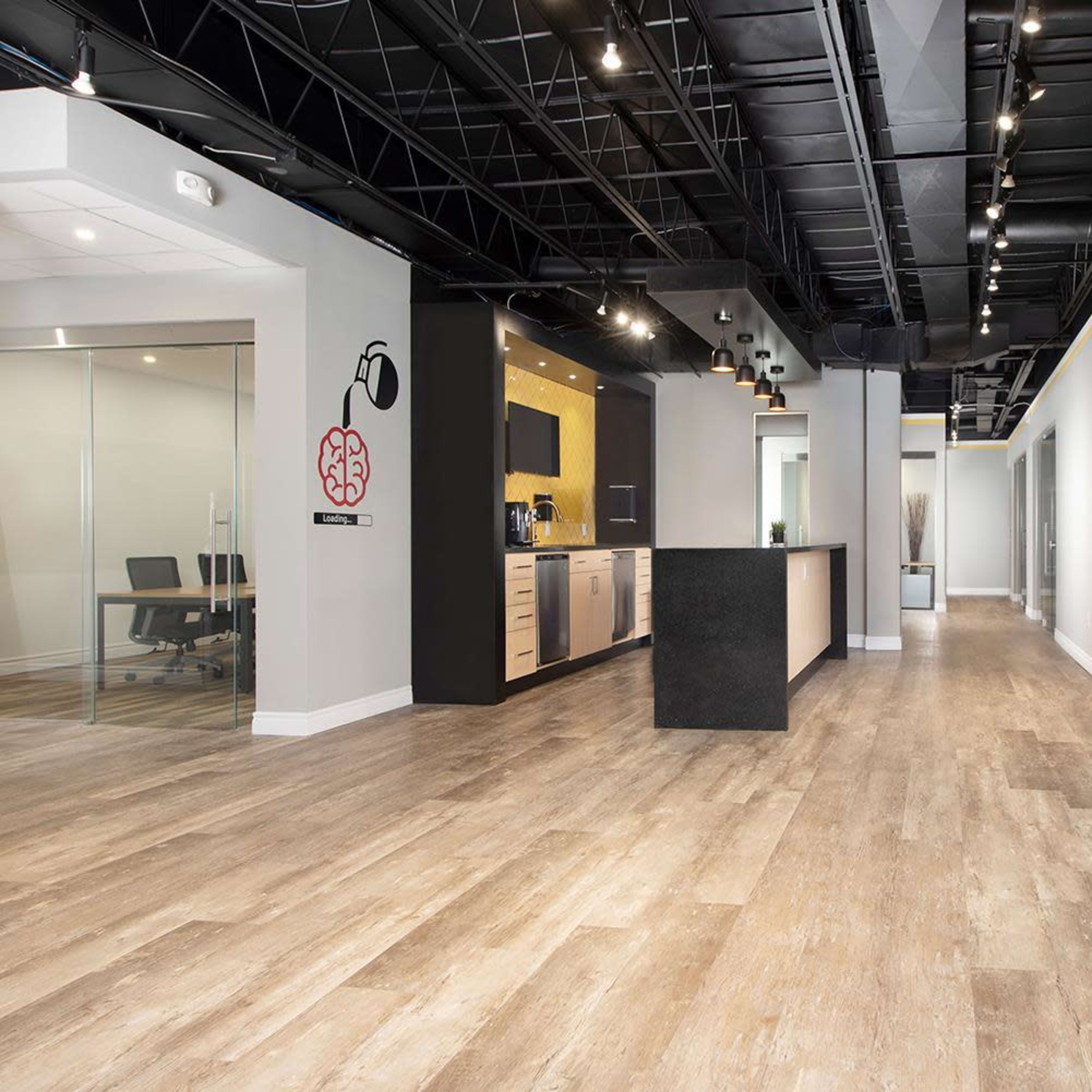 A modern office interior with a kitchen area on one side and glass-walled meeting rooms along the hallway.