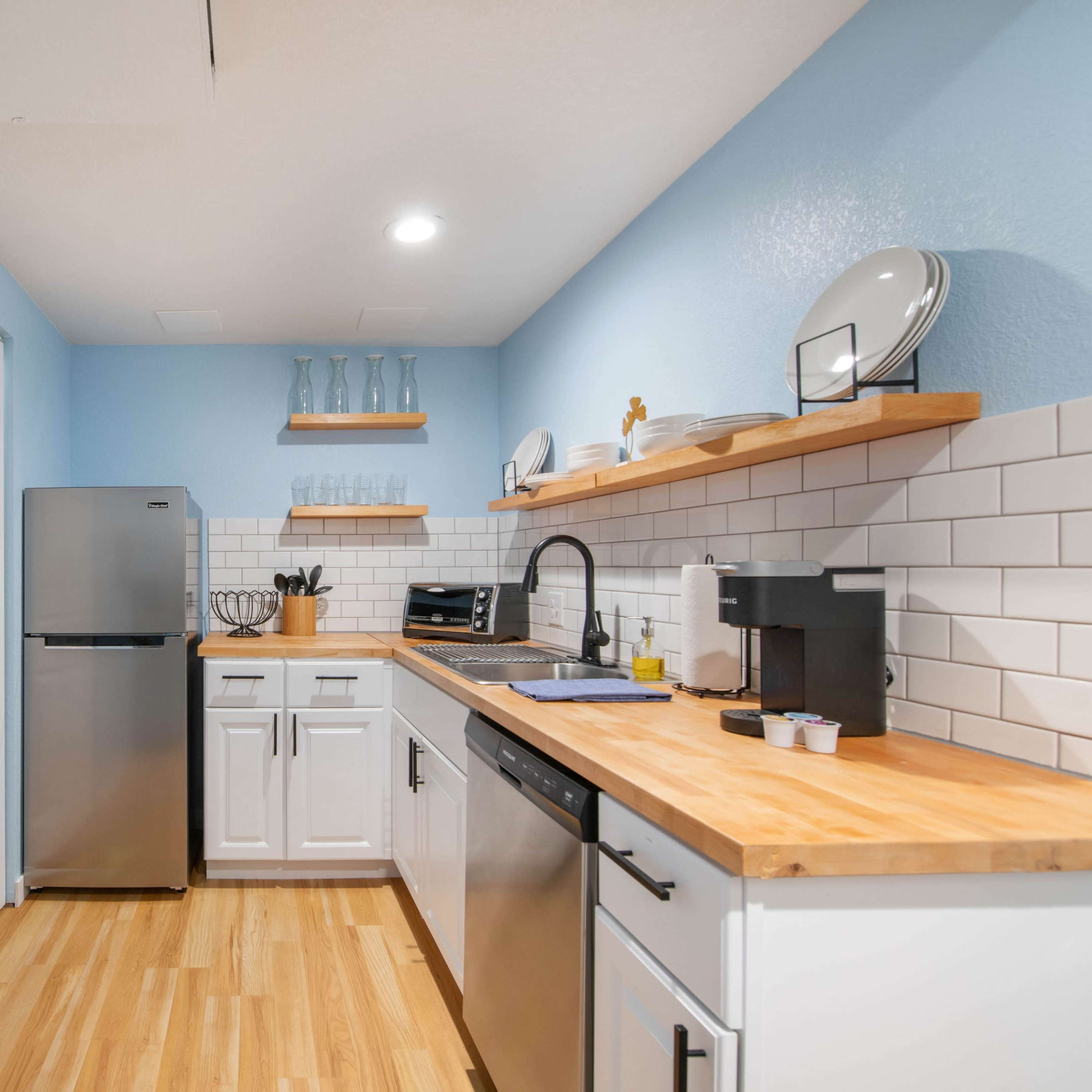 A modern kitchen features light blue walls, stainless steel appliances, white cabinetry, wooden countertops, and open shelving with decorative items.