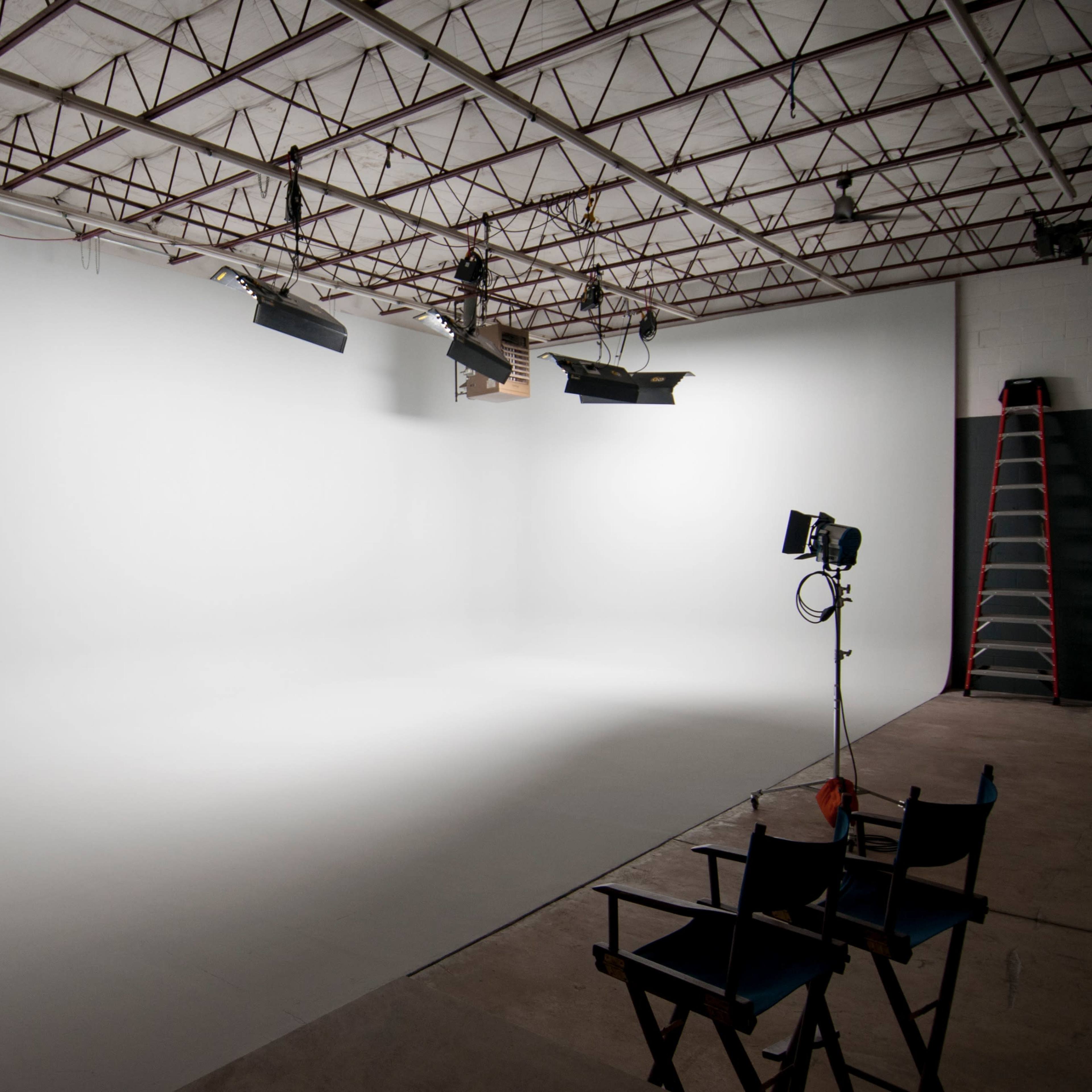 A photography studio features a seamless white backdrop, multiple lighting setups, and empty director's chairs.