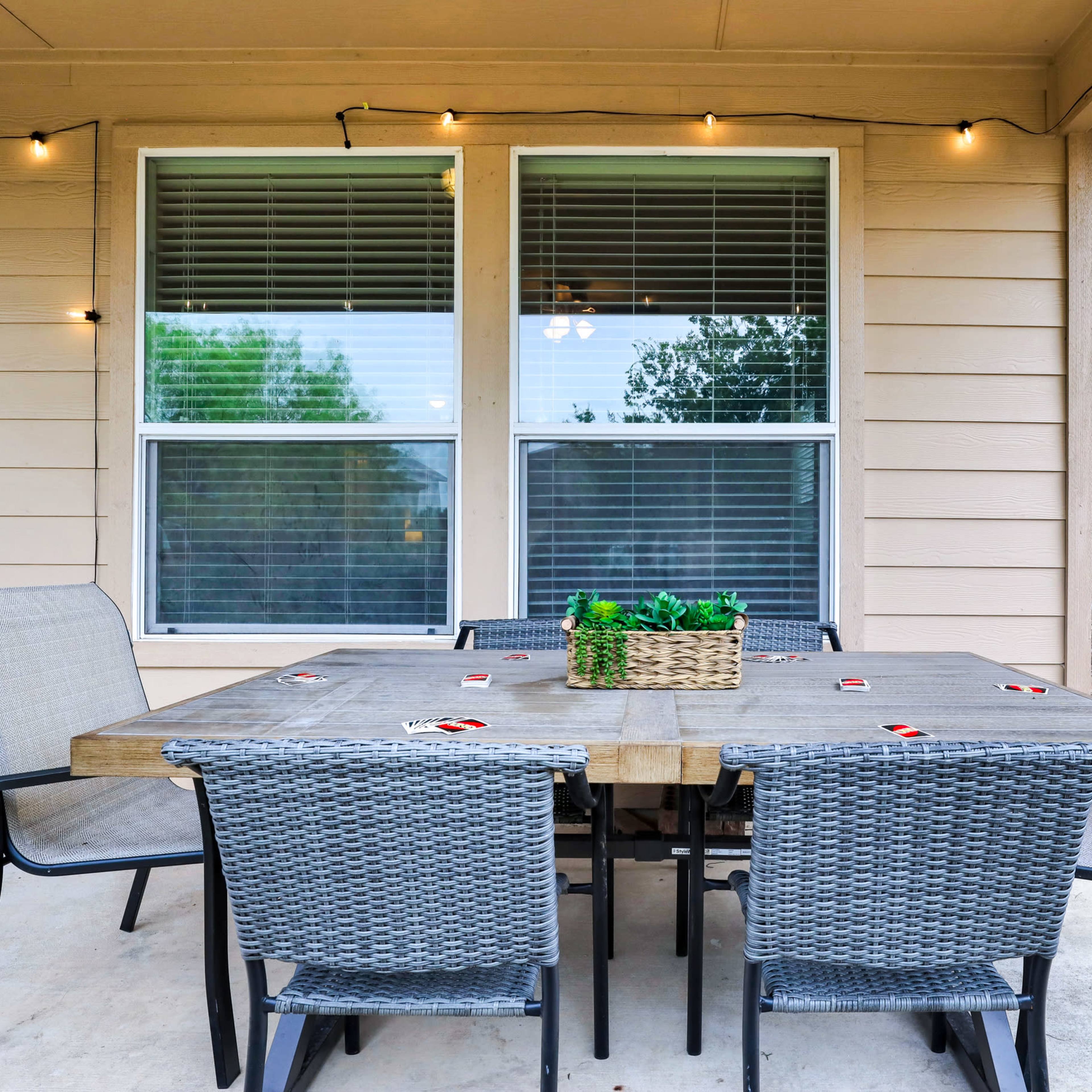 A patio dining set with a wooden table and six chairs is displayed outside a house, surrounded by string lights and windows.
