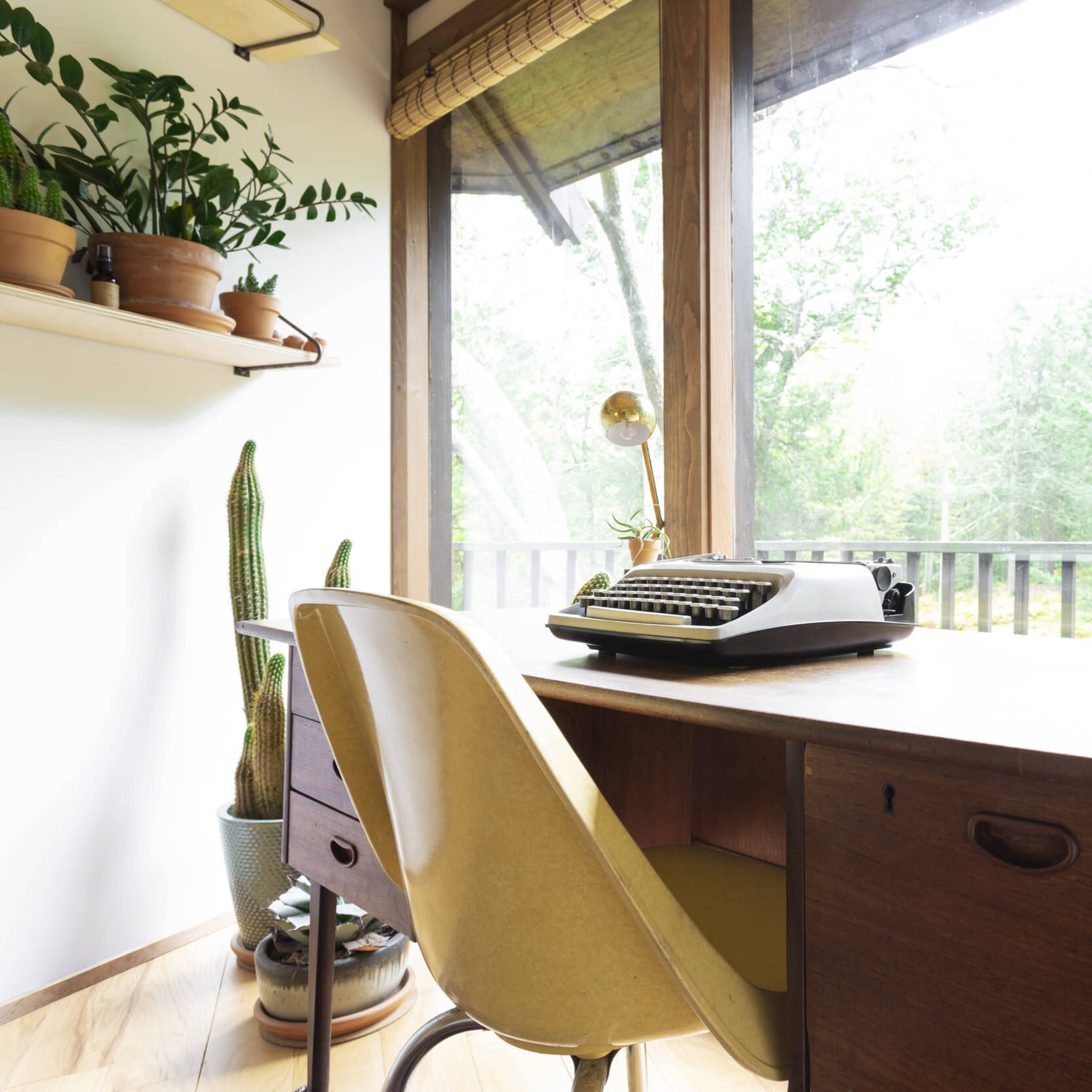 A wooden desk with a typewriter and a yellow chair is positioned beside large windows filled with greenery and potted plants.