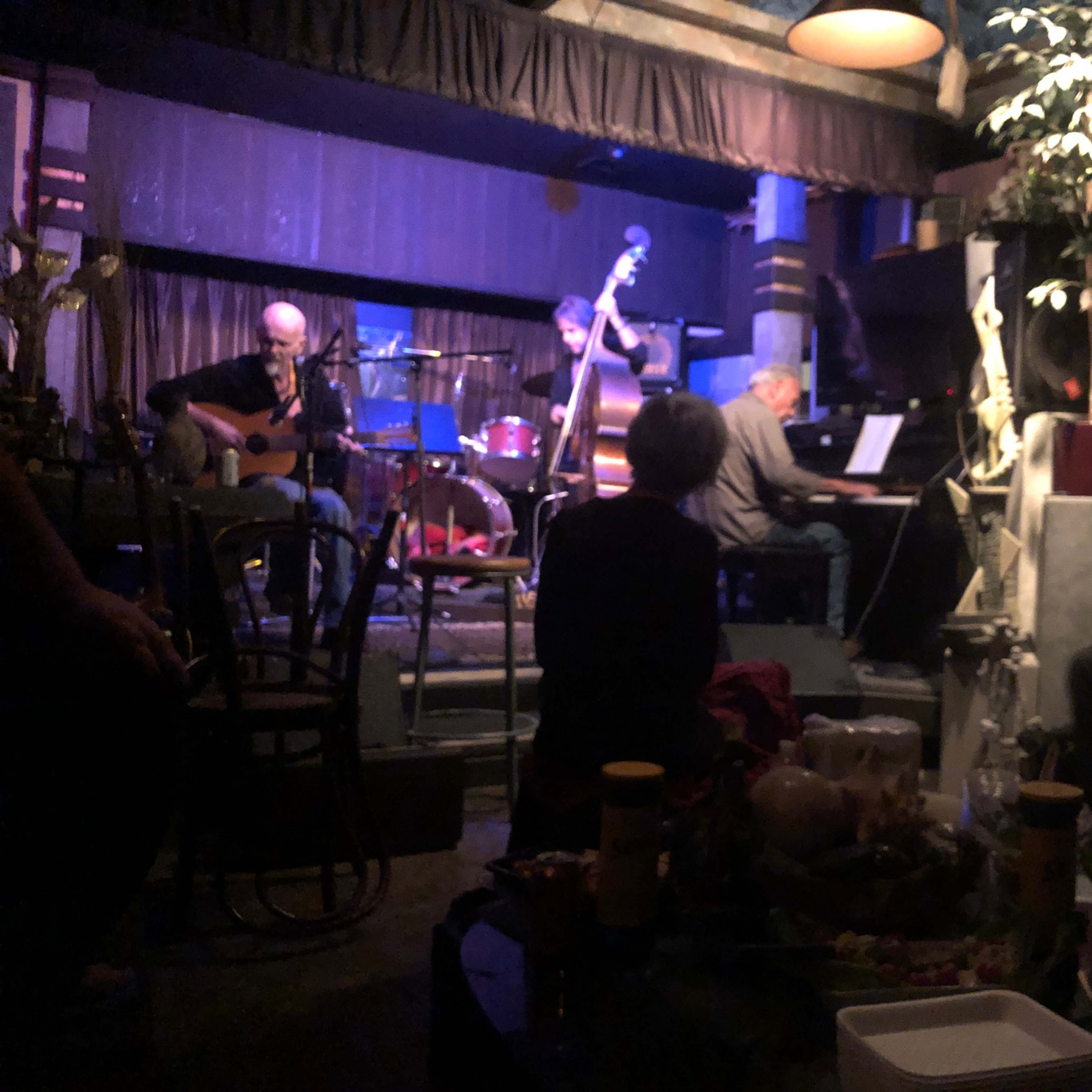 A small jazz band performs on a dimly lit stage in a cozy venue while an audience sits nearby.