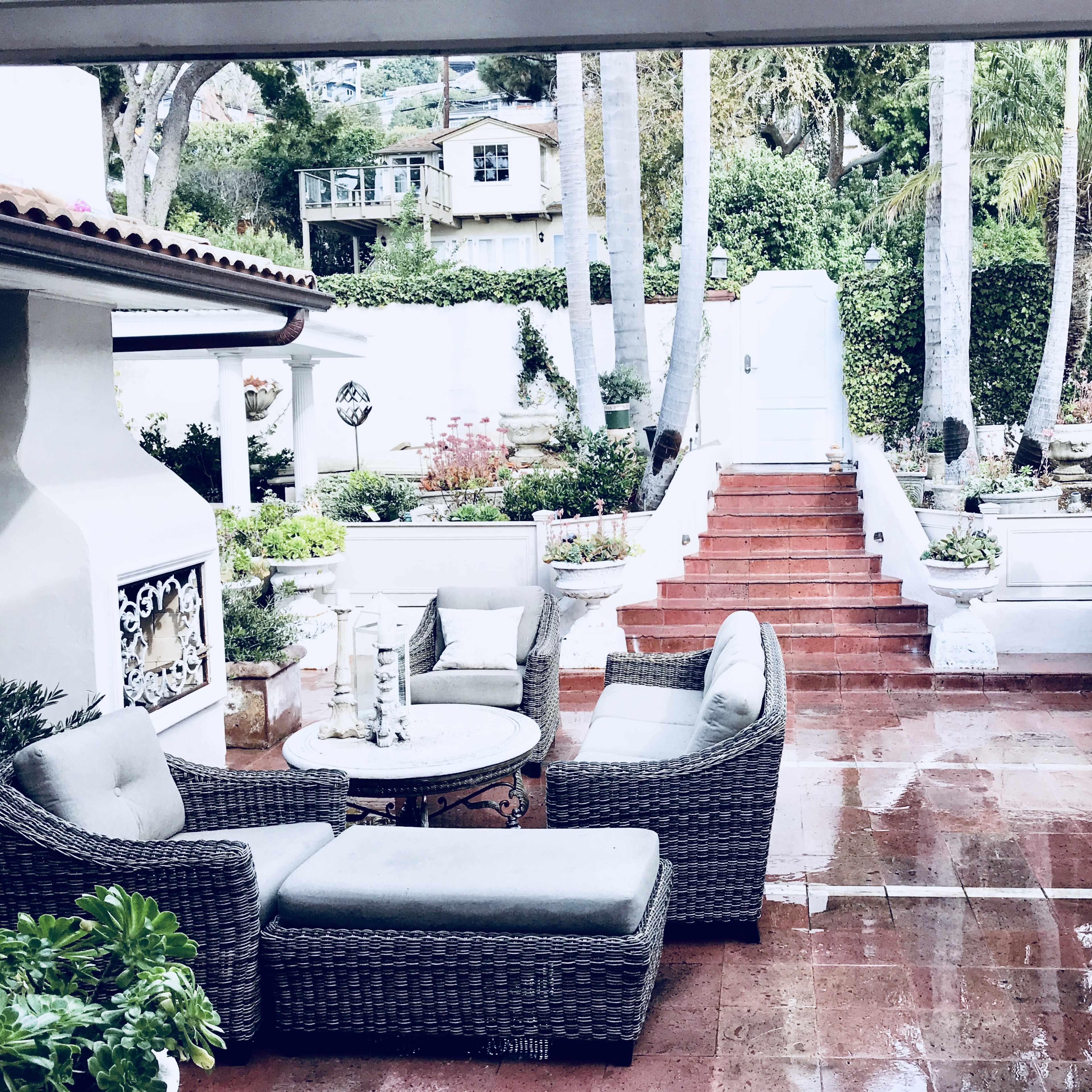 A landscaped patio area featuring wicker furniture, a stone fireplace, and a staircase leading to a white door surrounded by greenery.