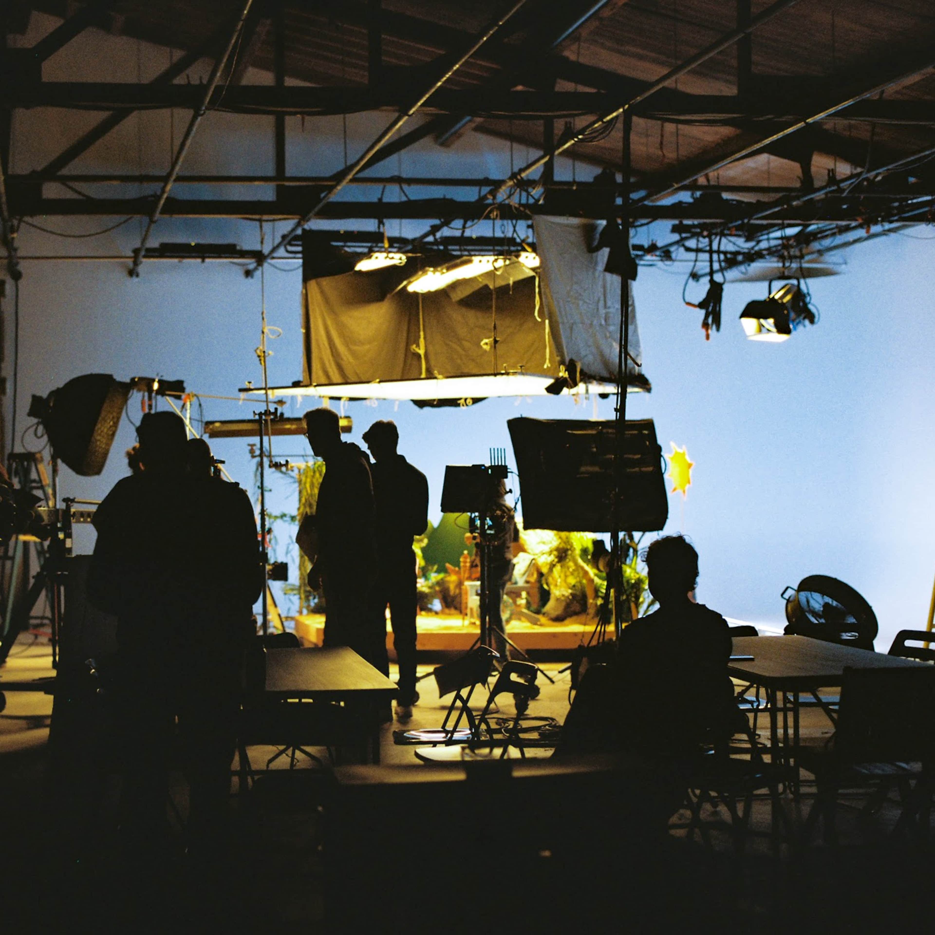 A production studio is filled with silhouettes of people setting up equipment under various lights and backdrops.