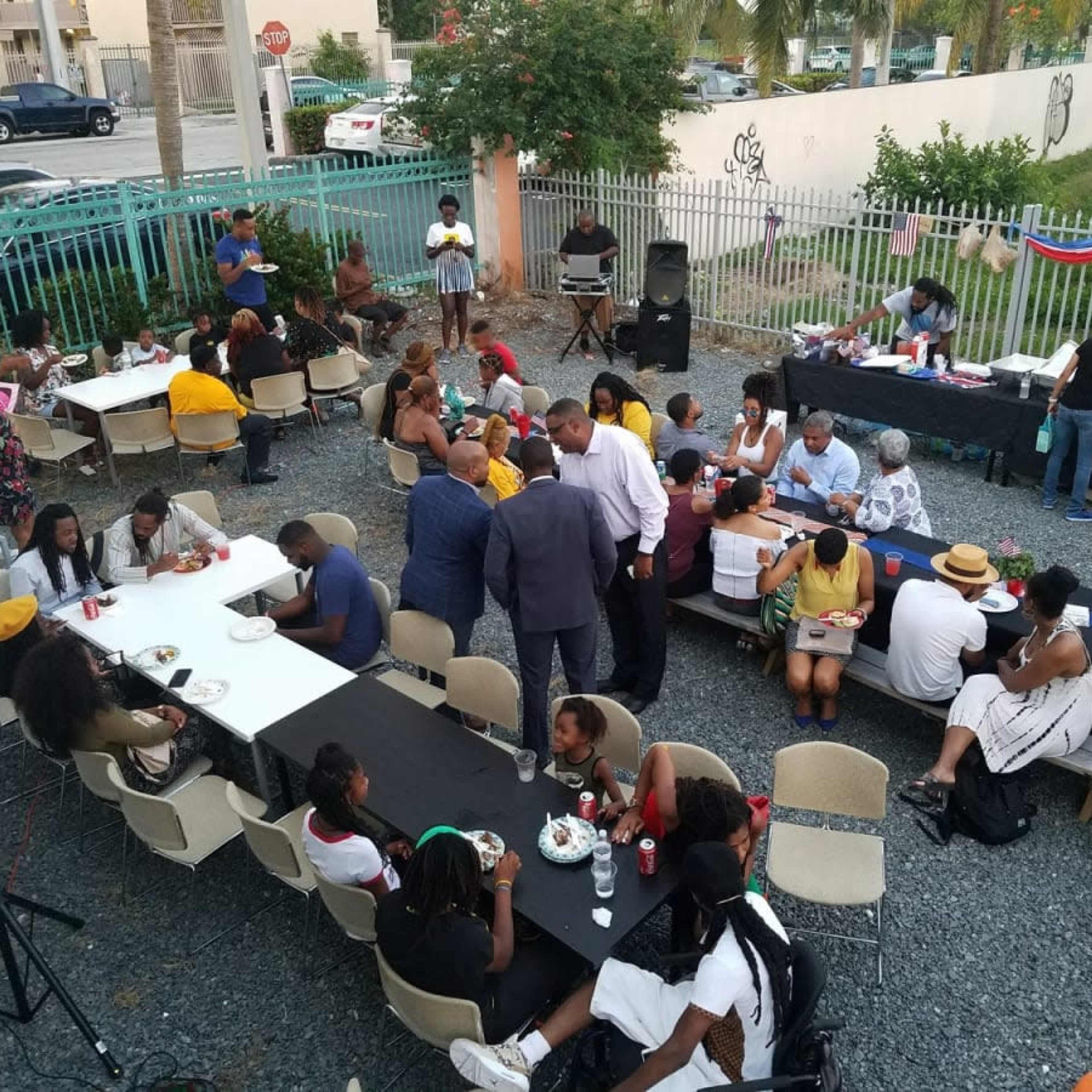A large group of people gathers around several tables outdoors for a social event, with food and drinks served in a community setting.