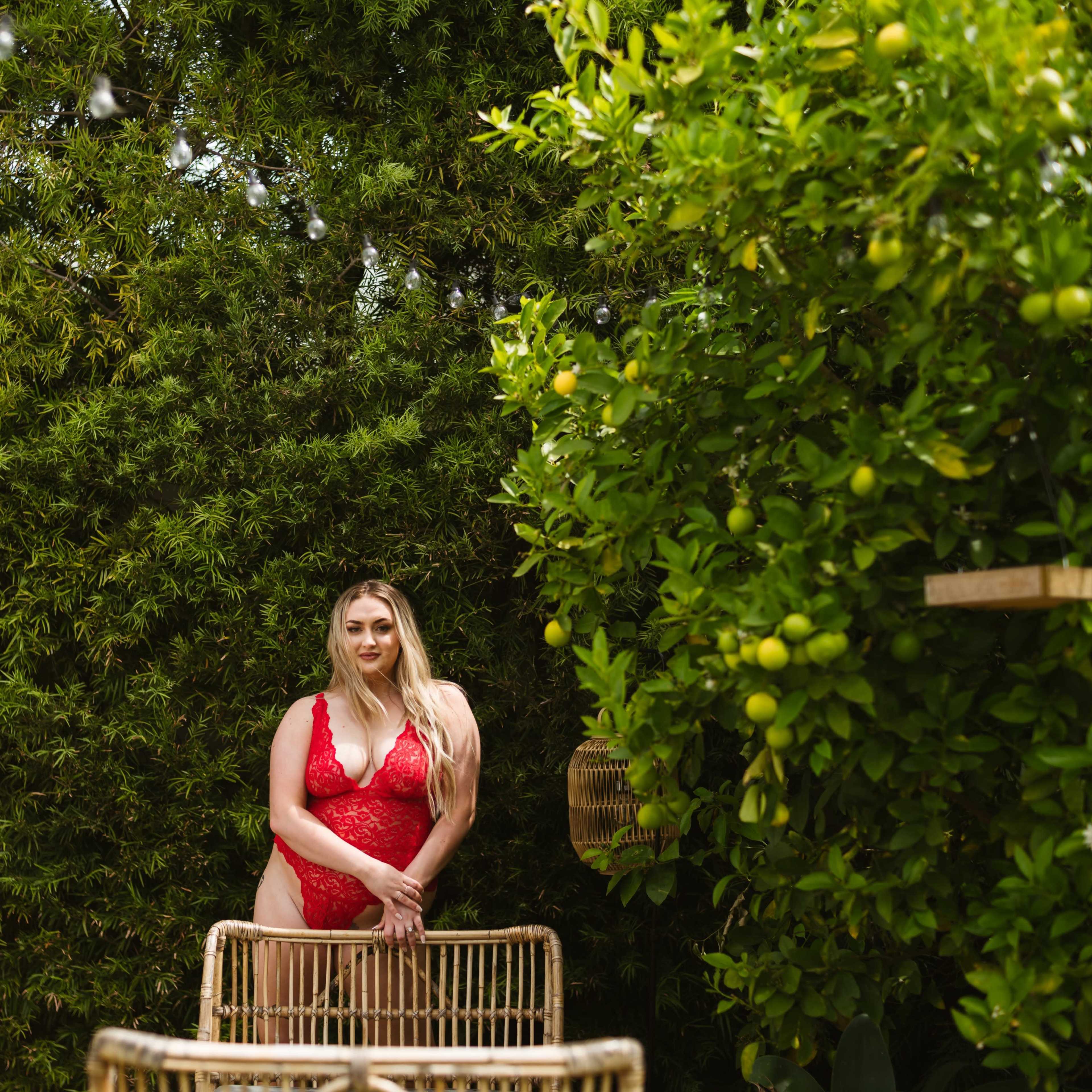 A woman in a red lingerie set stands beside a rattan chair in front of leafy greenery and a citrus tree.