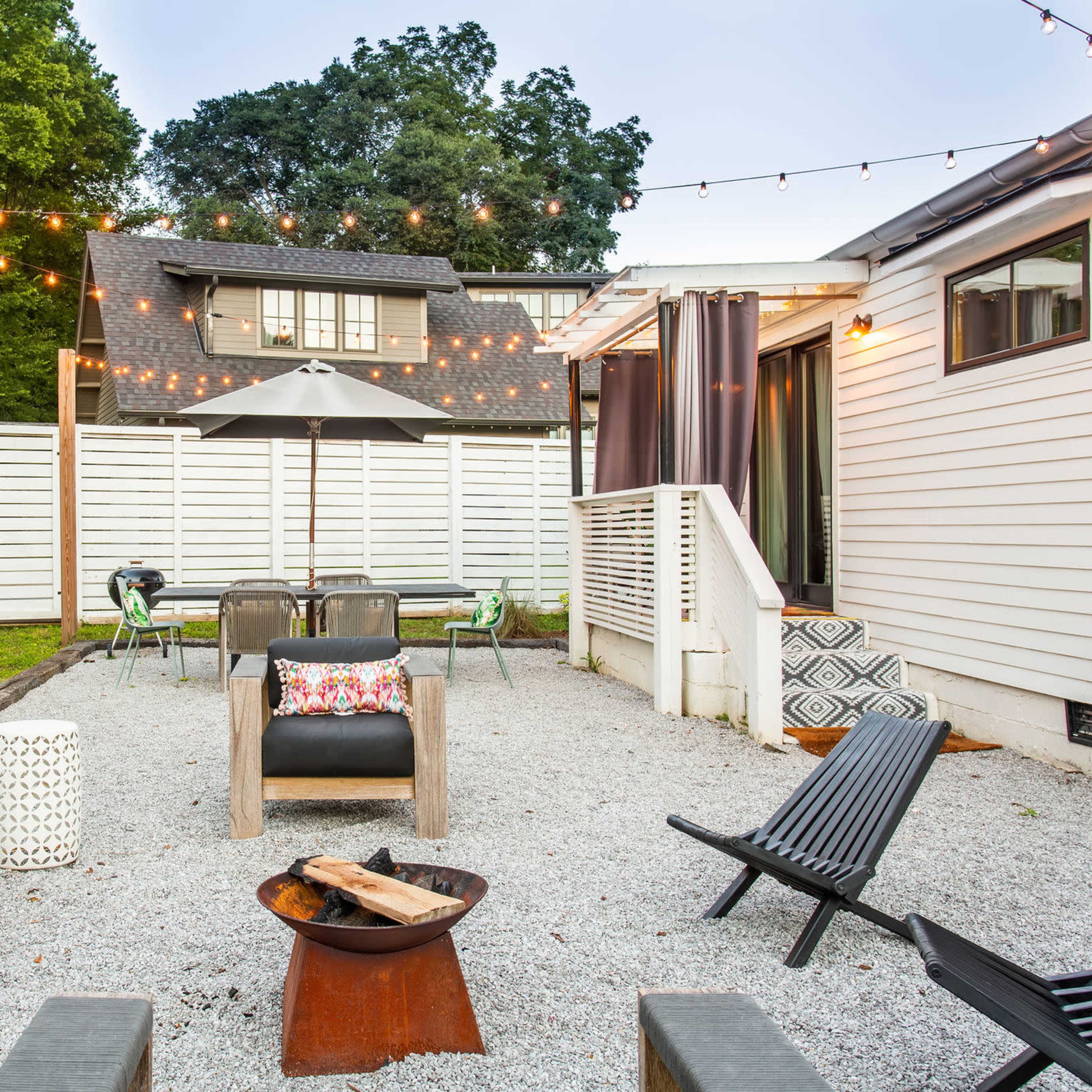 The scene shows a gravel backyard with seating arrangements, a fire pit, and string lights above an umbrella-covered table.