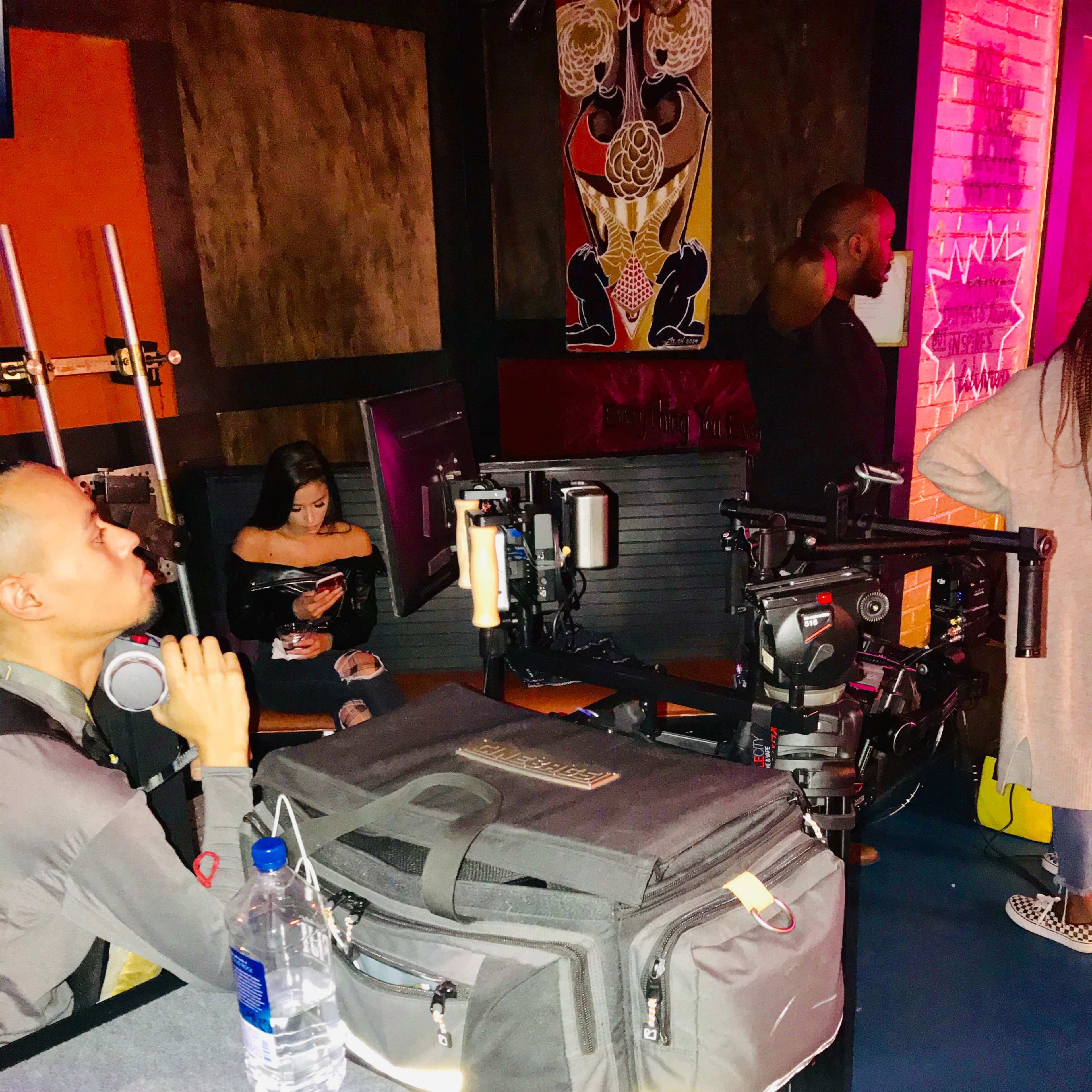 A person sits at a table with a water bottle and camera equipment while others are visible in the background.
