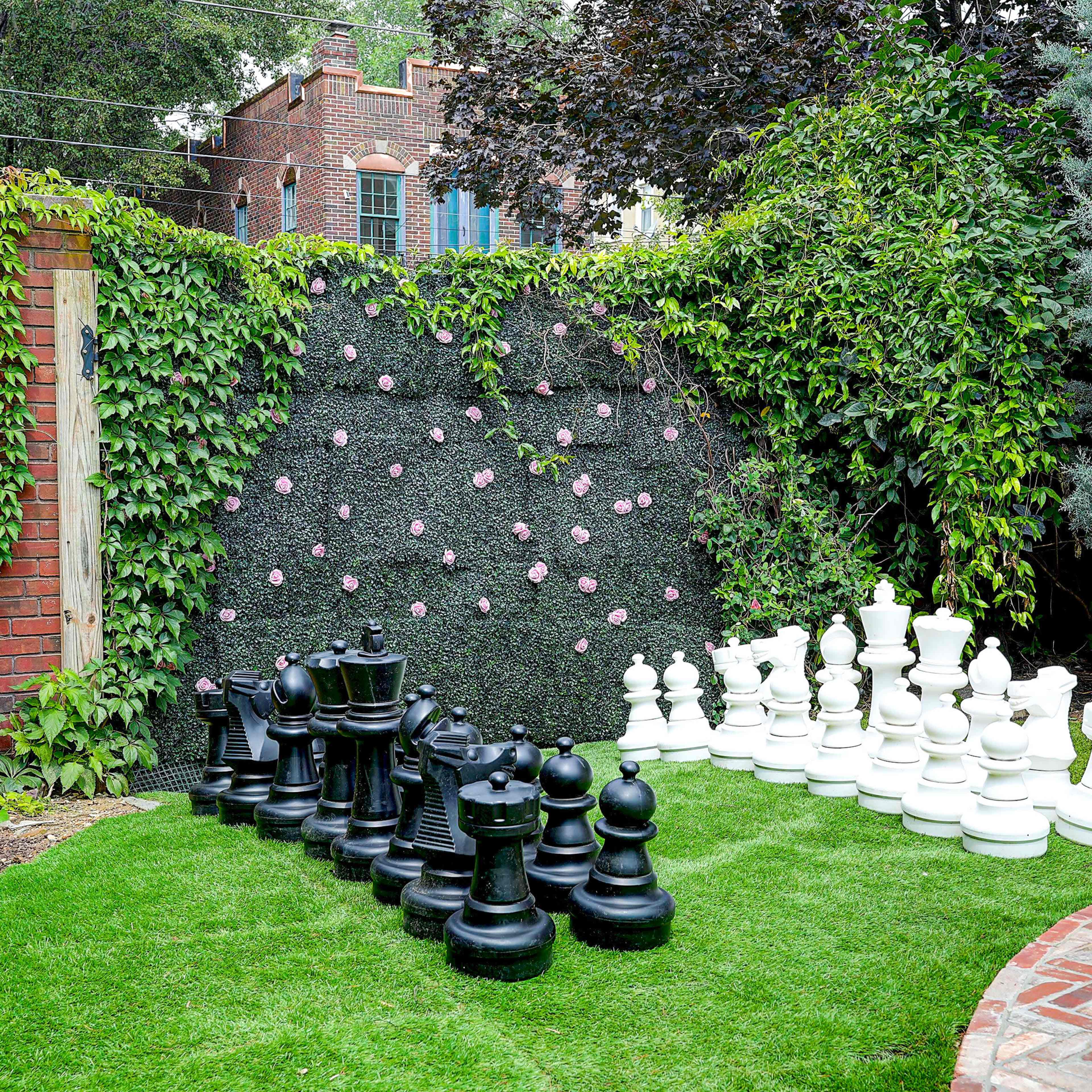 A large outdoor chess set made of black and white pieces is arranged on a grassy area, surrounded by greenery and a brick walkway.