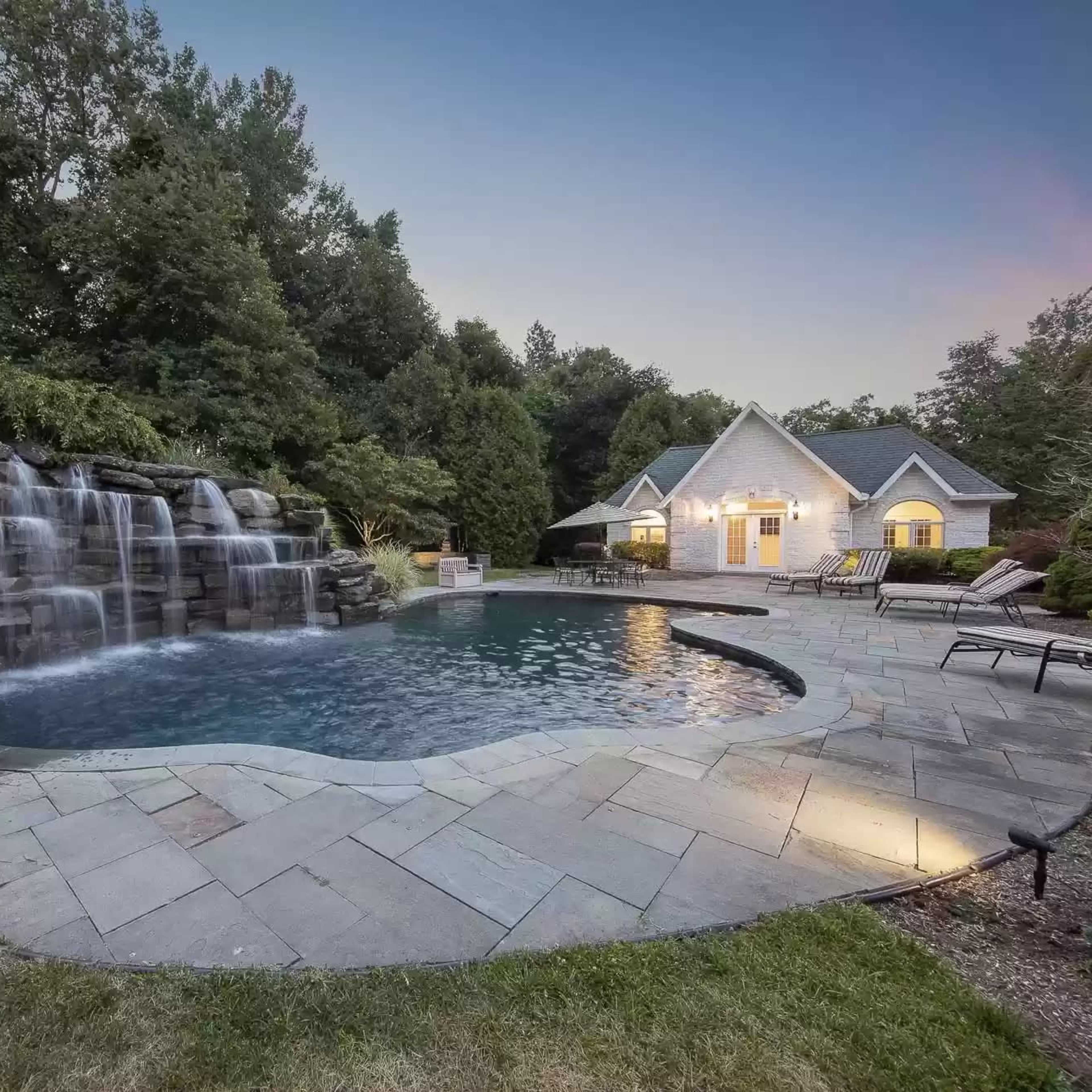 A landscaped backyard featuring a swimming pool with a waterfall surrounded by stone tiles and a house in the background.