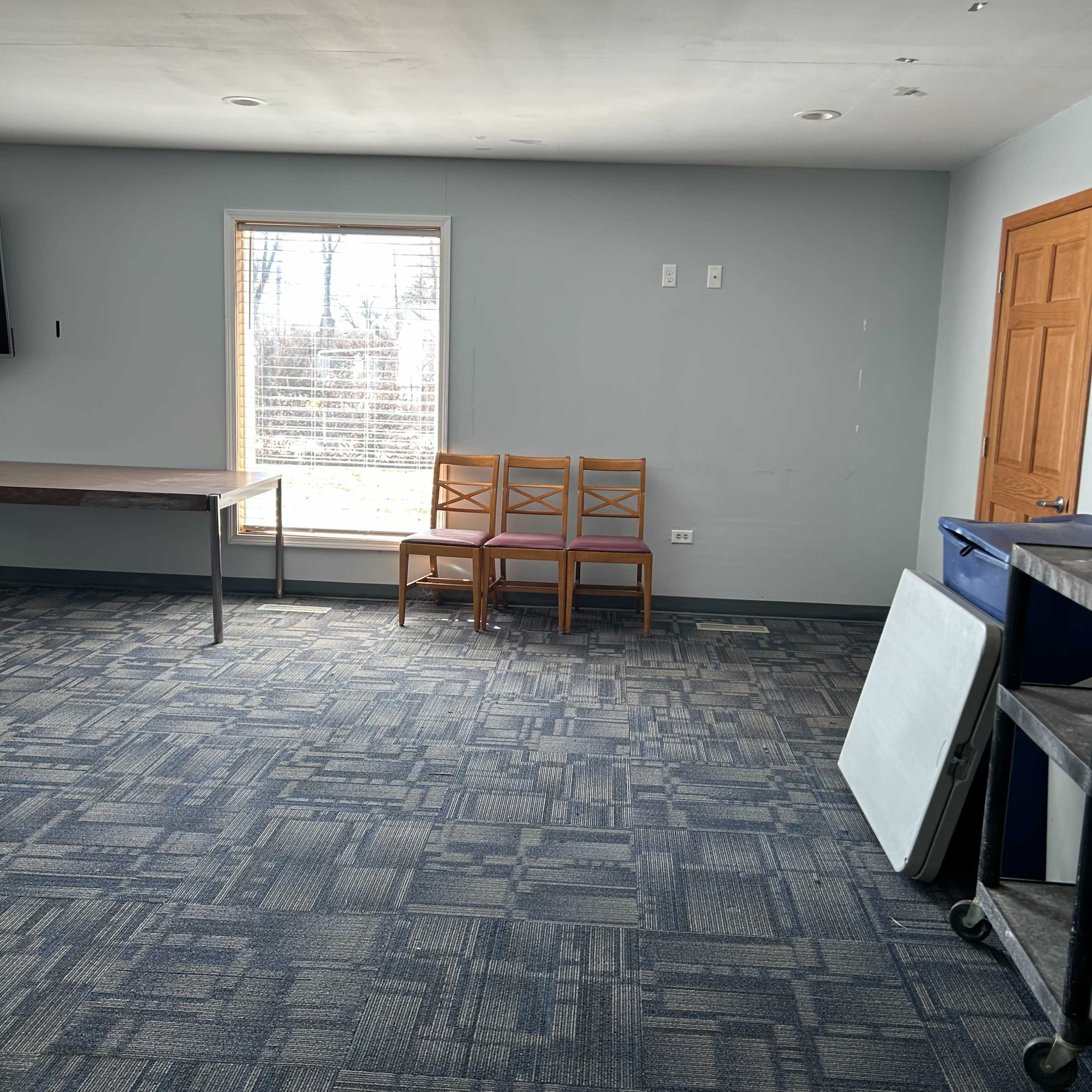 A small, empty room features a window, three wooden chairs against the wall, and a couple of tables positioned around the space.