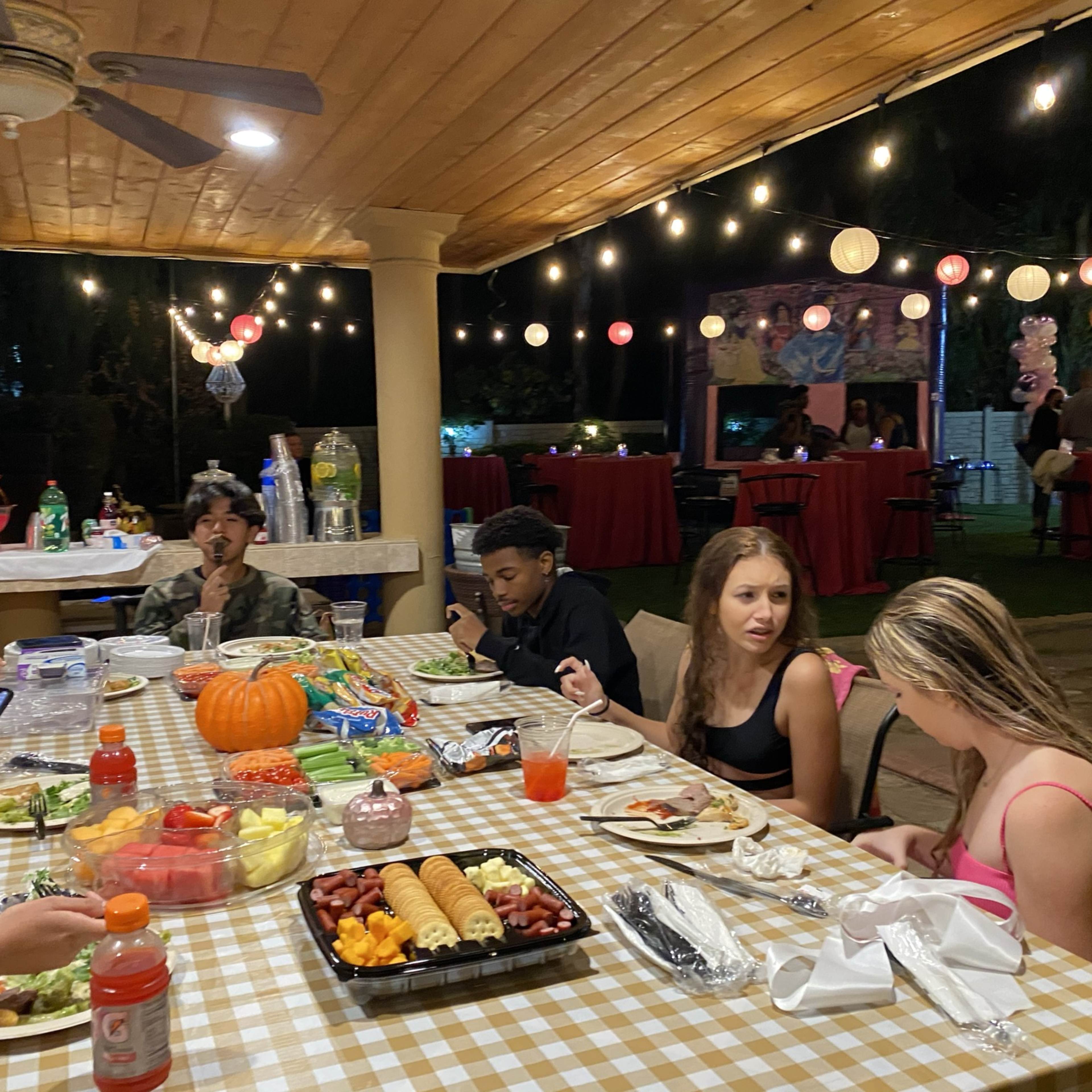 A group of people enjoy a meal at a long table adorned with various dishes, while string lights illuminate the outdoor setting.