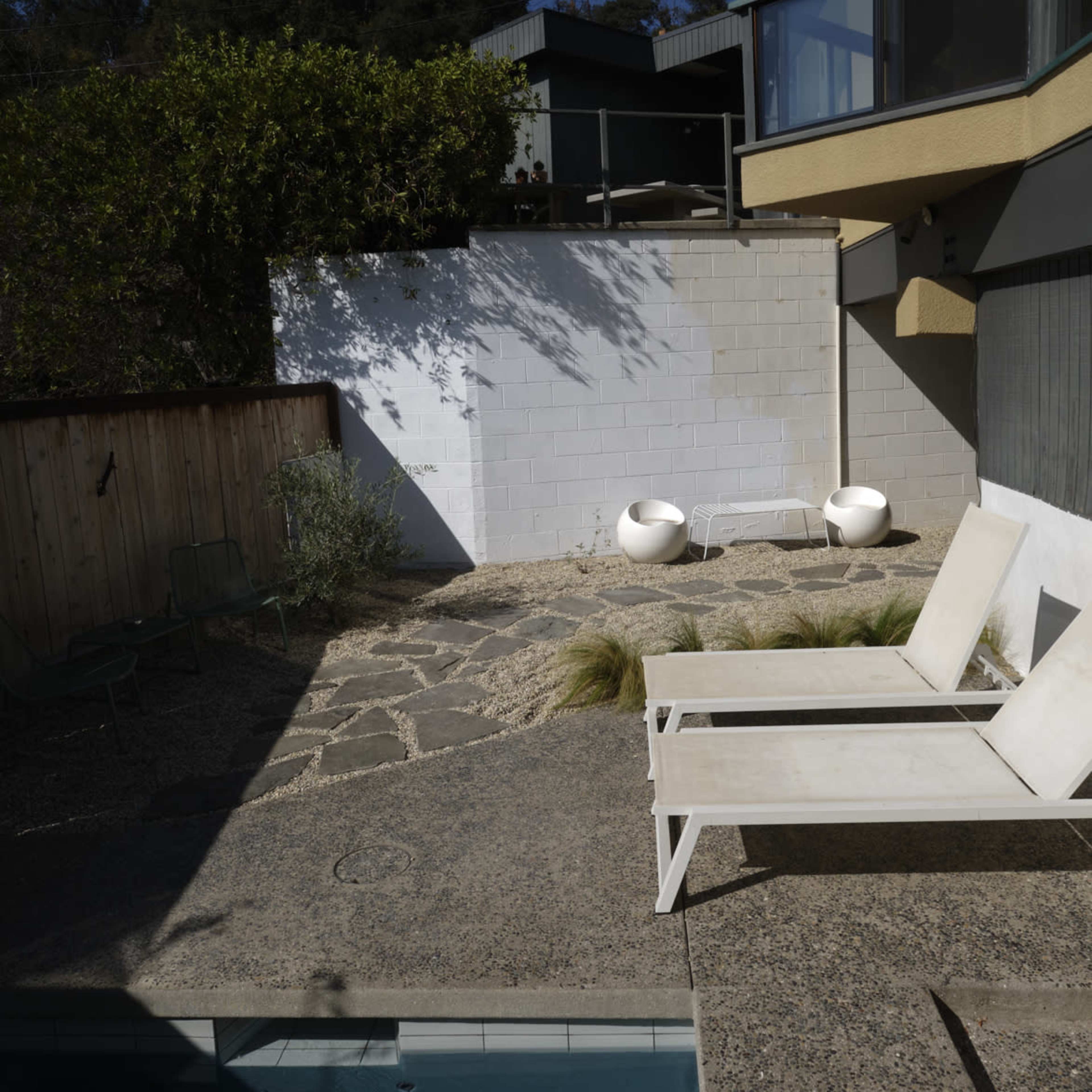 A sunlit patio area with two white lounge chairs beside a pool, adjacent to a stone pathway and a wall with greenery above.