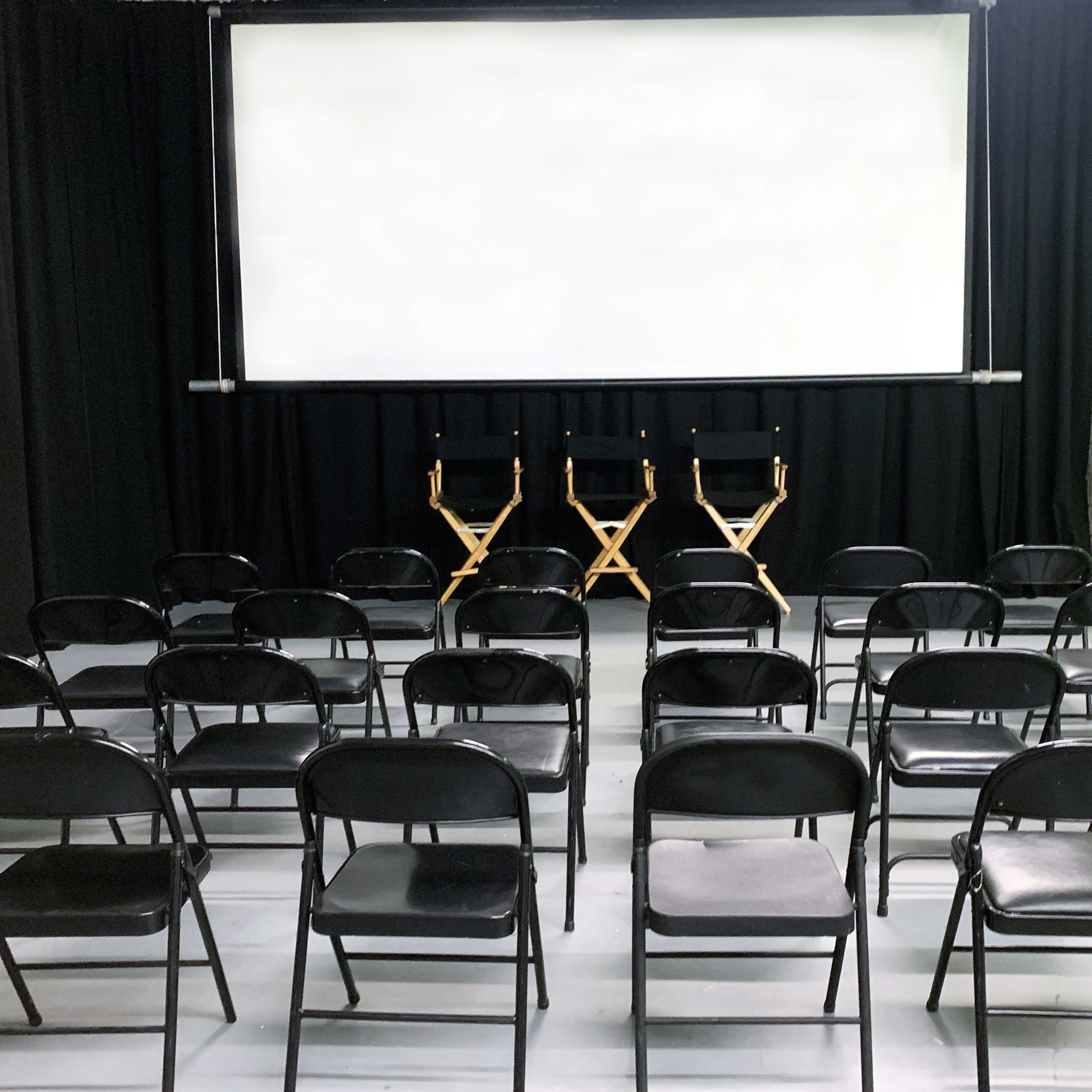 A screening room with black folding chairs arranged in rows facing a blank screen and two director's chairs on stage.