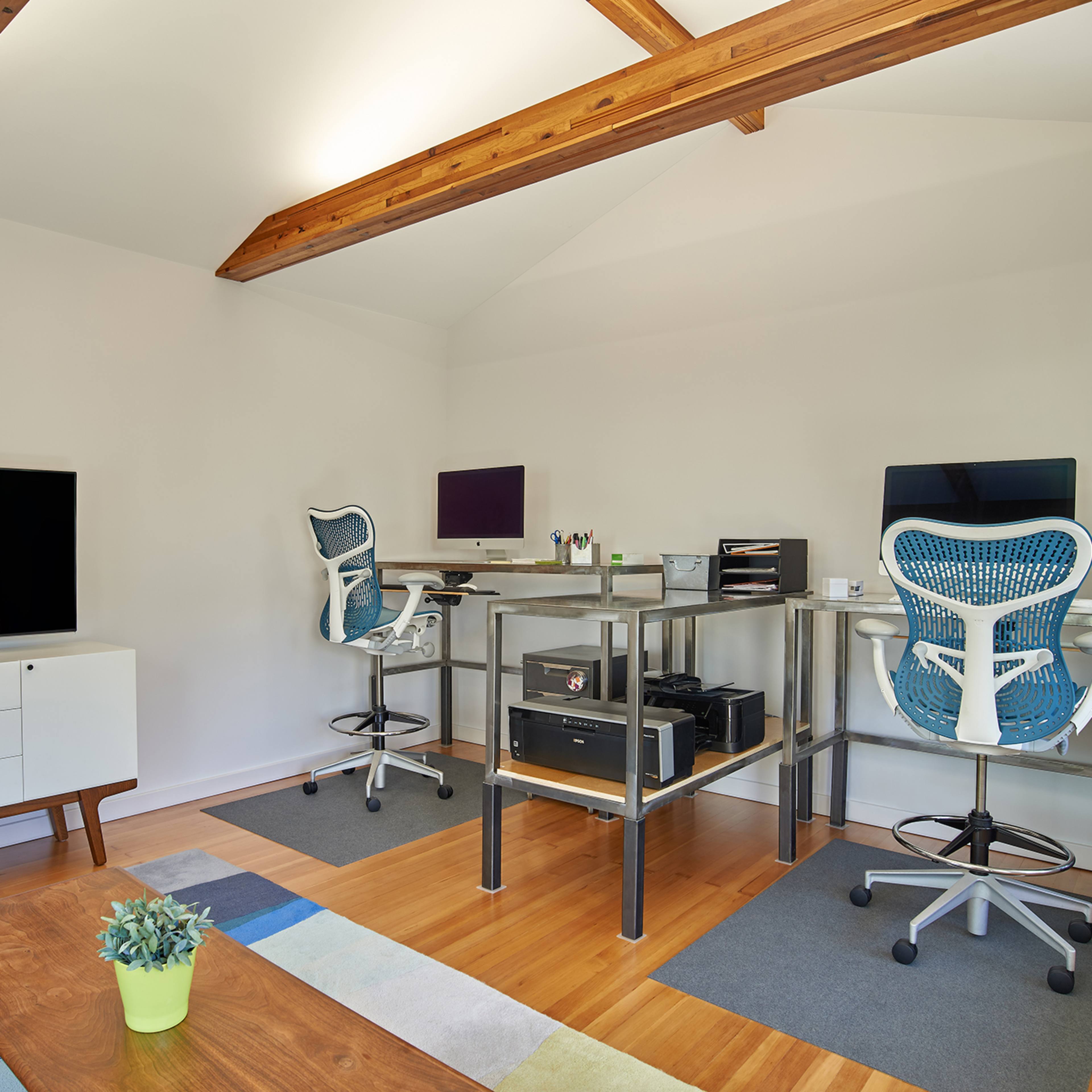 A modern home office setup featuring two desks with ergonomic chairs, a wall-mounted television, and light hardwood flooring.