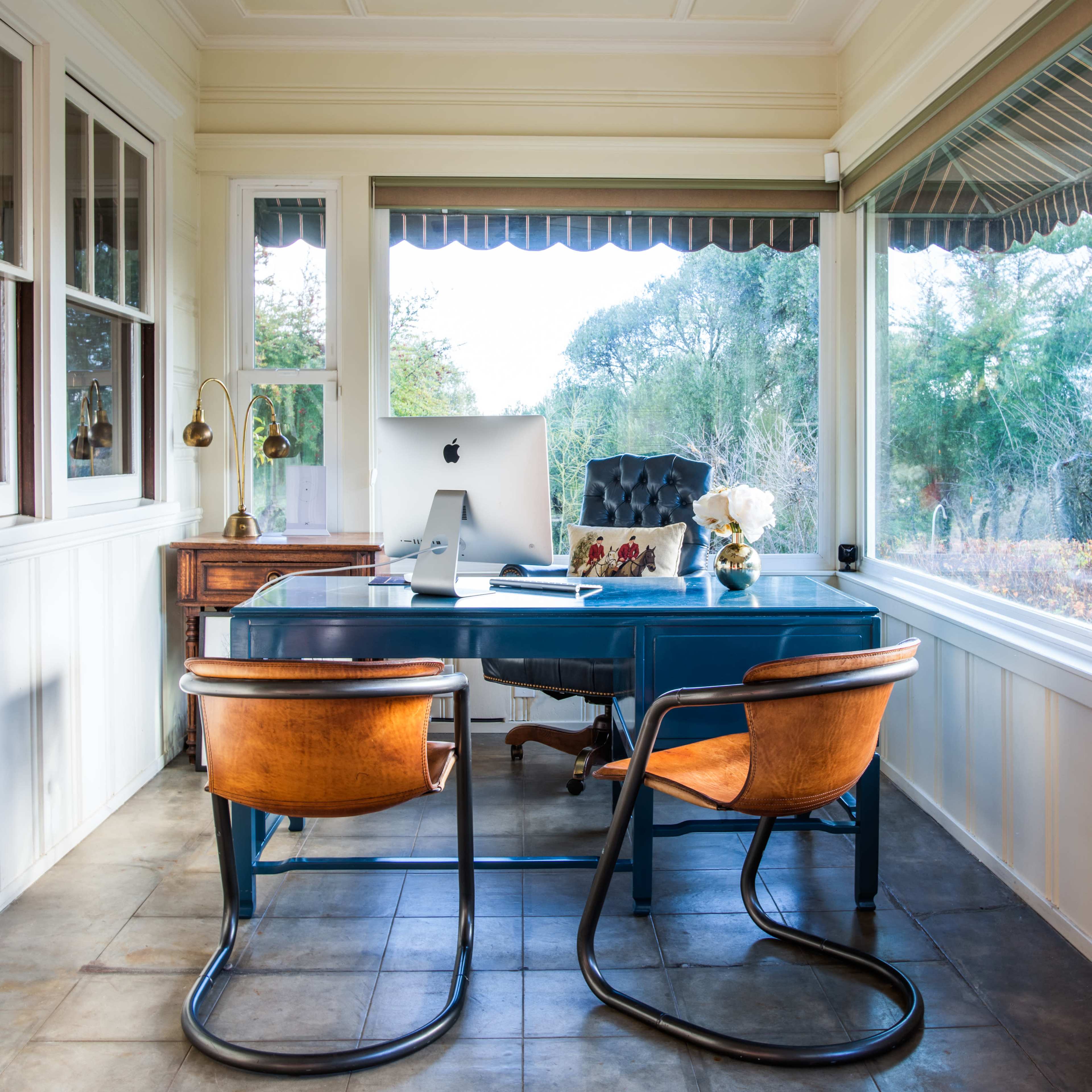 A stylish home office features a blue desk with an iMac, two curved wooden chairs, and large windows showcasing a view of greenery outside.