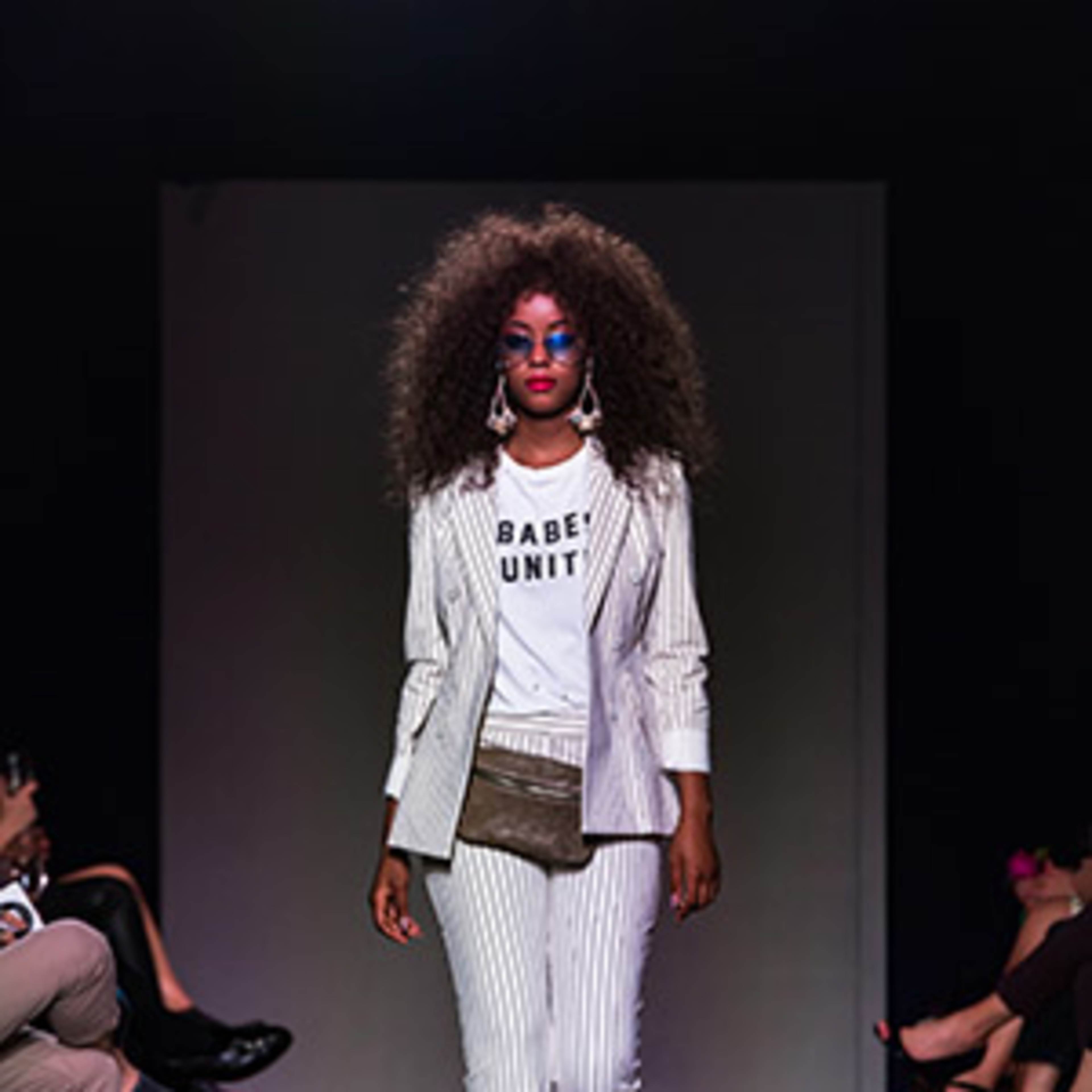 A model walks down a runway wearing a striped suit over a graphic t-shirt, with an audience seated on either side.