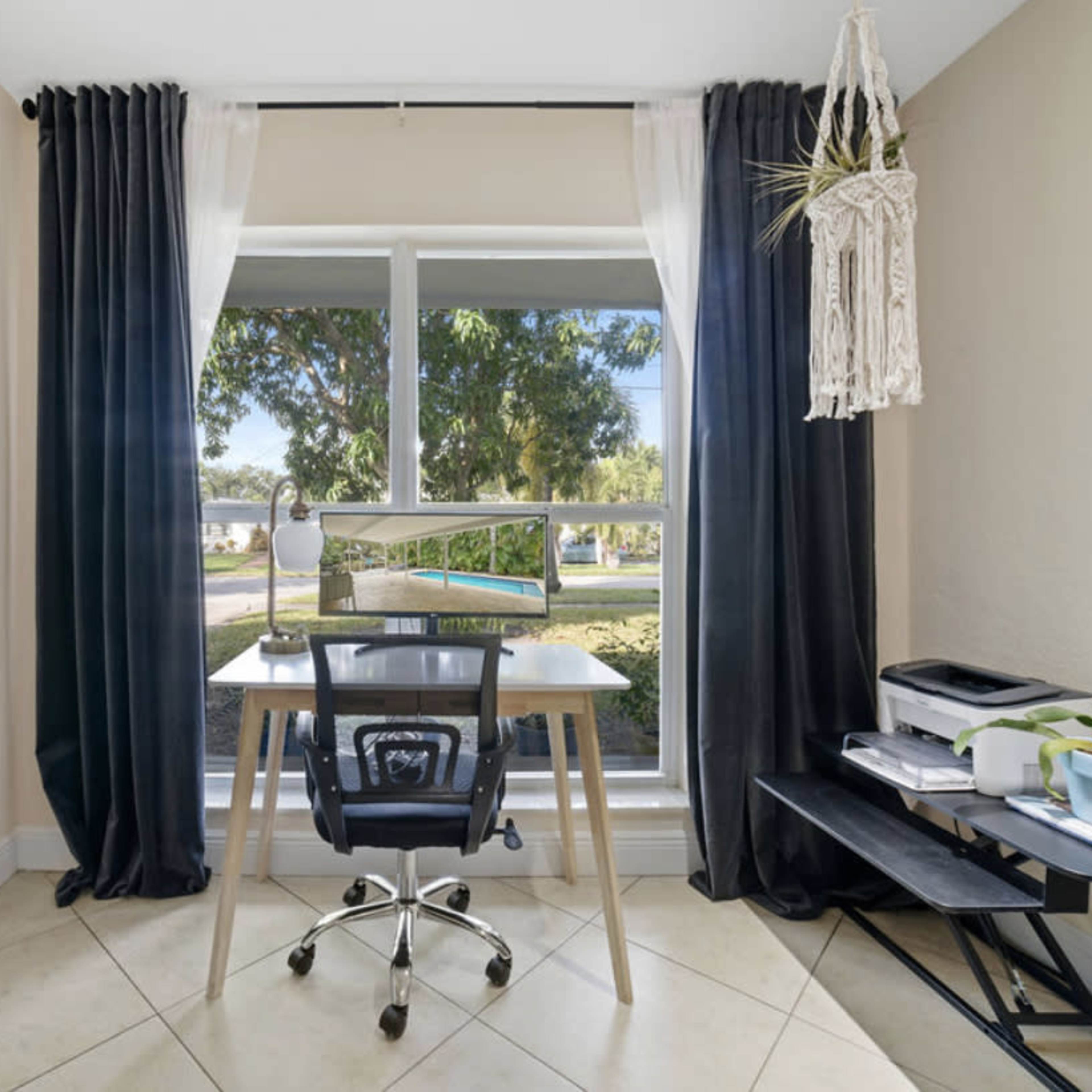 A workspace is situated next to a large window, featuring a desk with a chair, a monitor, and a small shelf with plants.