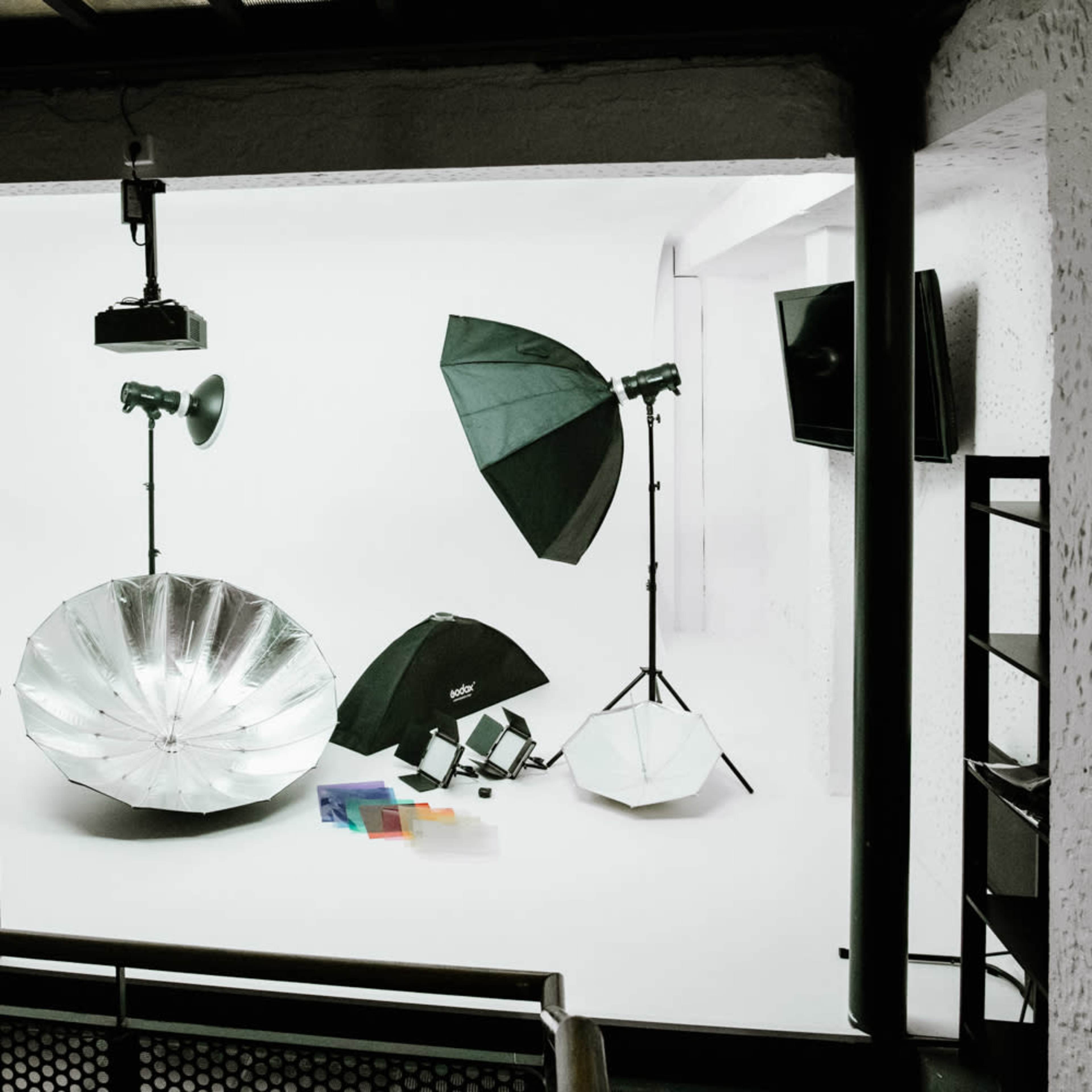 A photography studio setup is shown with various lighting equipment, including umbrellas and softboxes, arranged against a white backdrop.