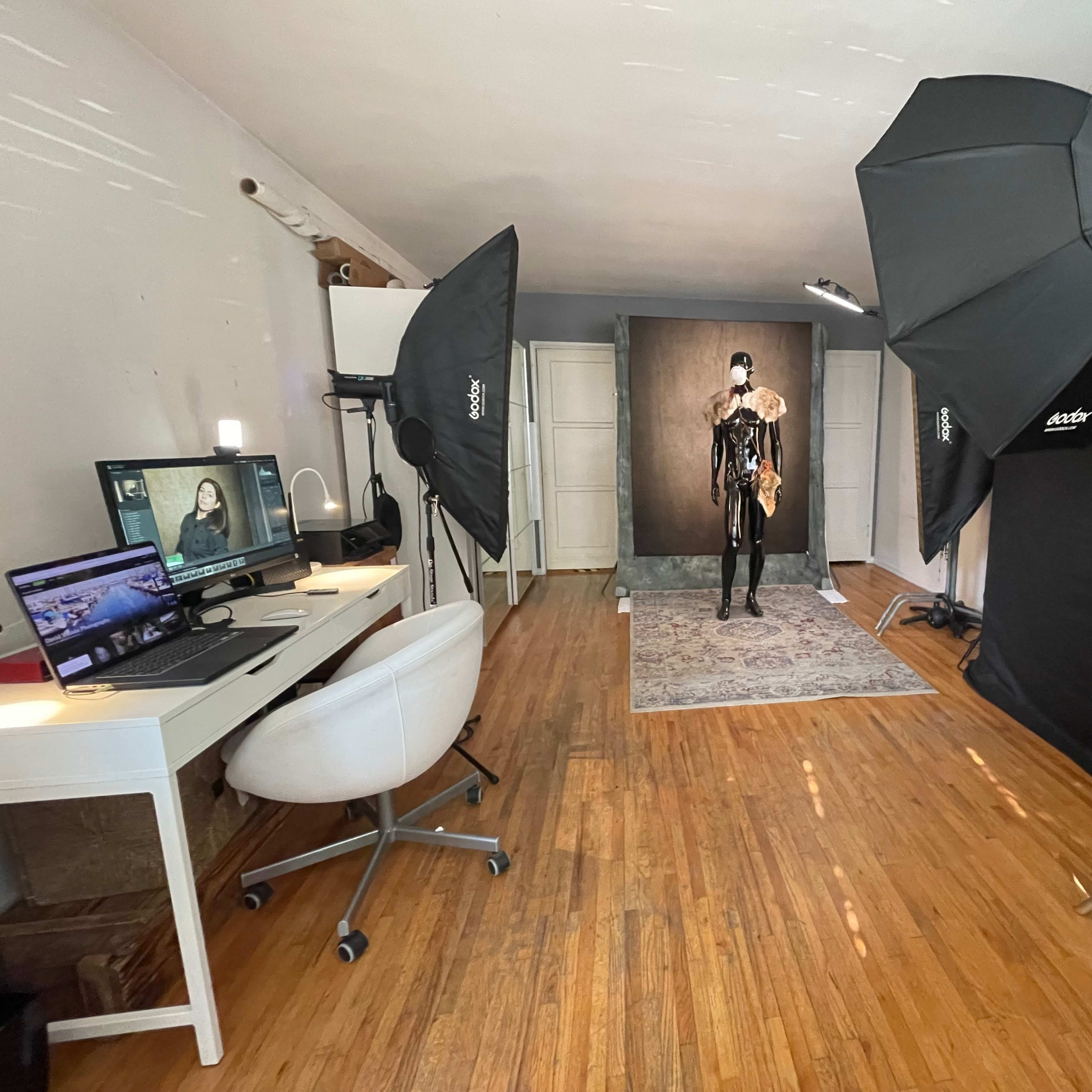 A photography studio with a model in a black outfit standing on a rug, surrounded by lighting equipment and a computer workstation.