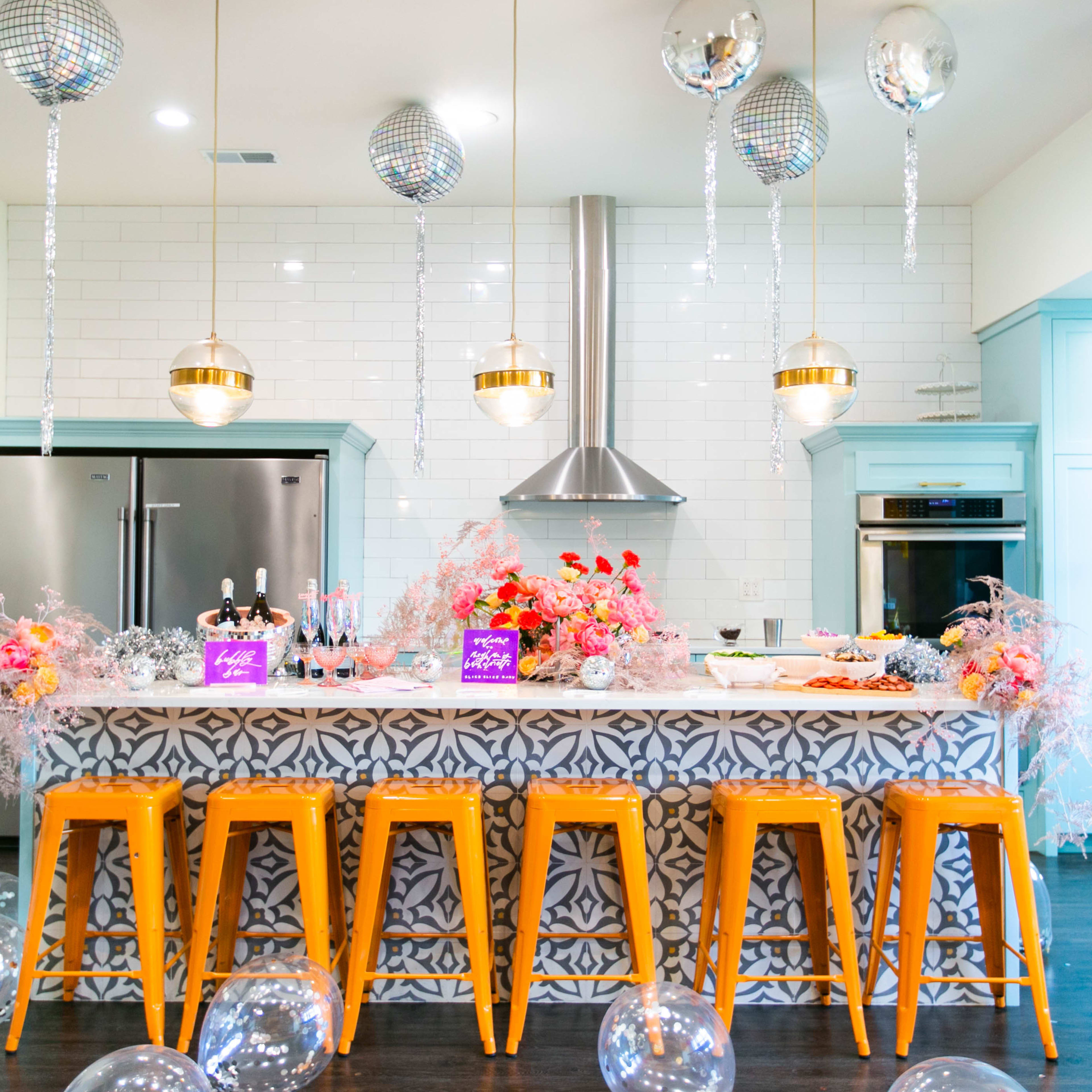 A modern kitchen features a vibrant bar with orange stools, a marble countertop adorned with floral decorations, and silver disco balls hanging from the ceiling.